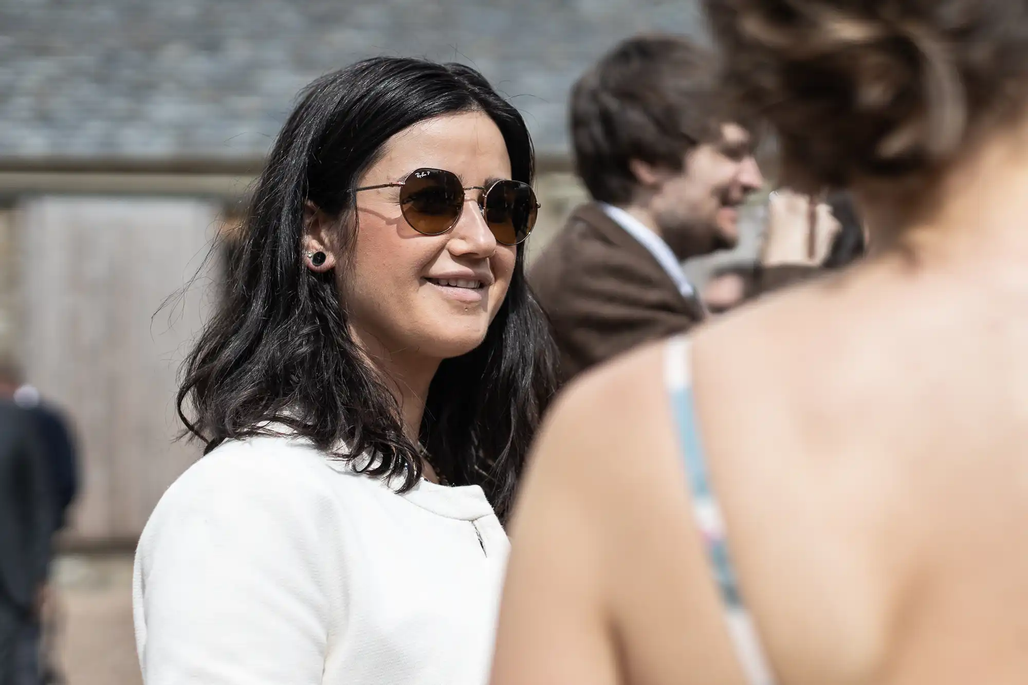 A woman in sunglasses and white top smiling and conversing at a sunny outdoor social event.