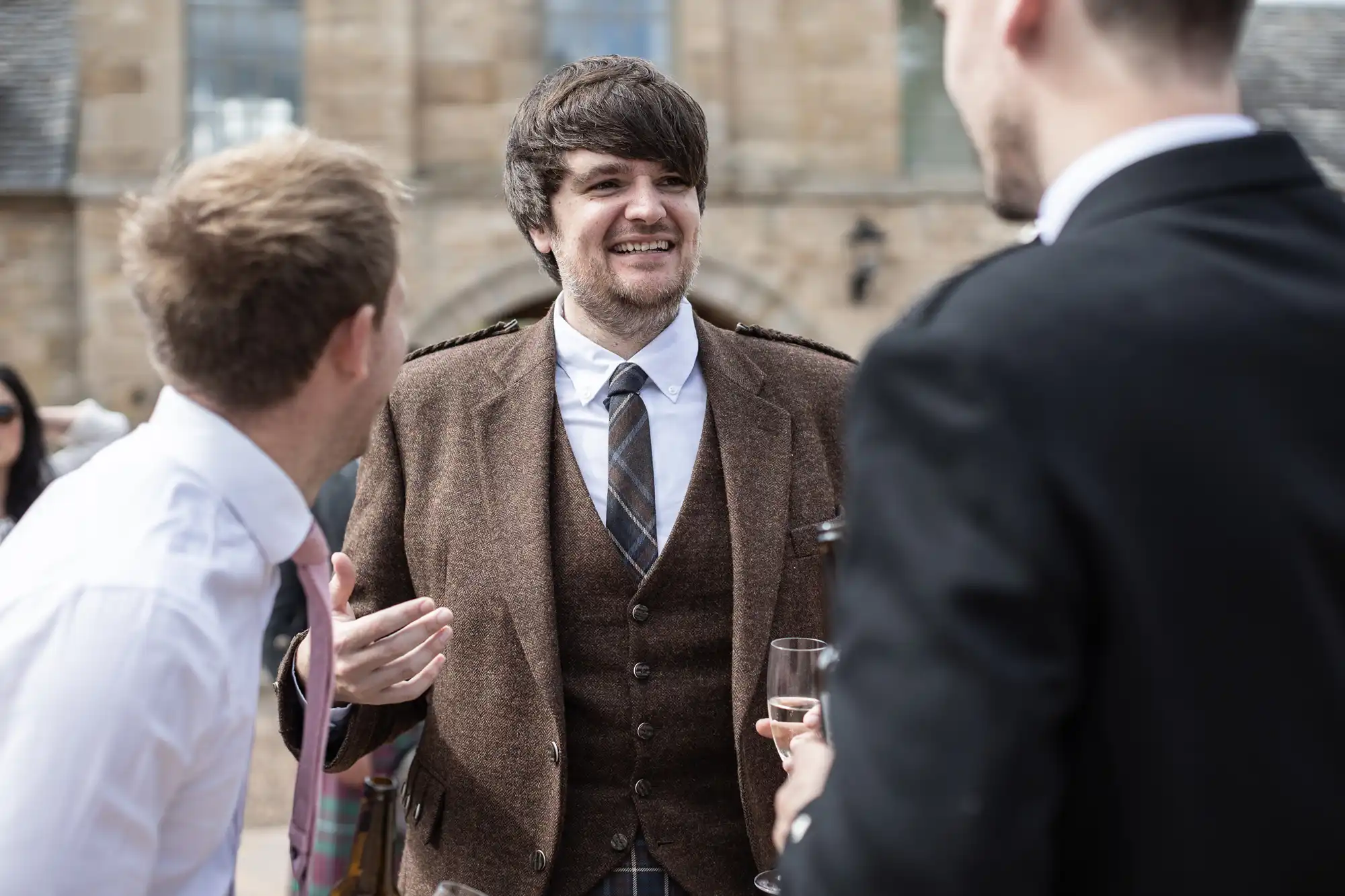 A man in a brown tweed jacket and tie laughs while chatting with two other men at an outdoor gathering.