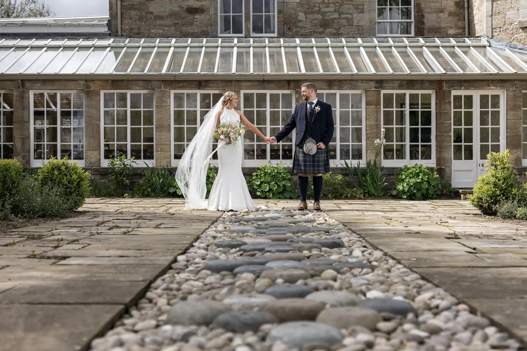 A bride in a white dress and a groom in a kilt holding hands in front of a stone building with large windows and a glass roof.