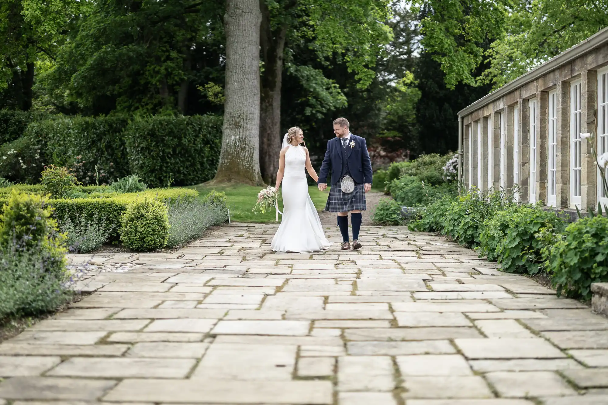 A bride and groom walk hand in hand along a stone pathway in a garden, with lush greenery and a classical structure in the background.