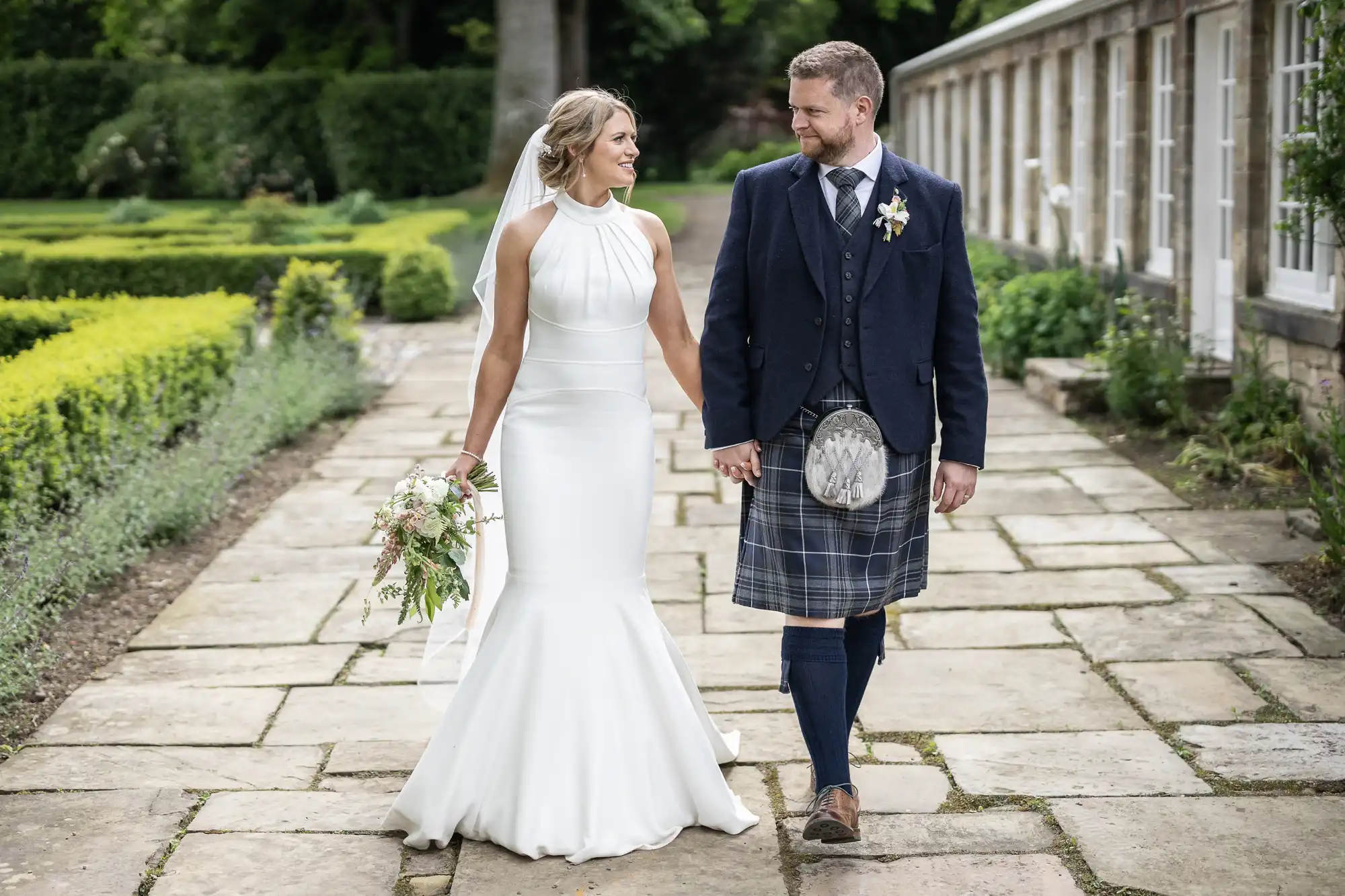 A bride in a white dress and a groom in a kilt holding hands and walking along a garden path, smiling at each other on their Rosebery Steading wedding day.