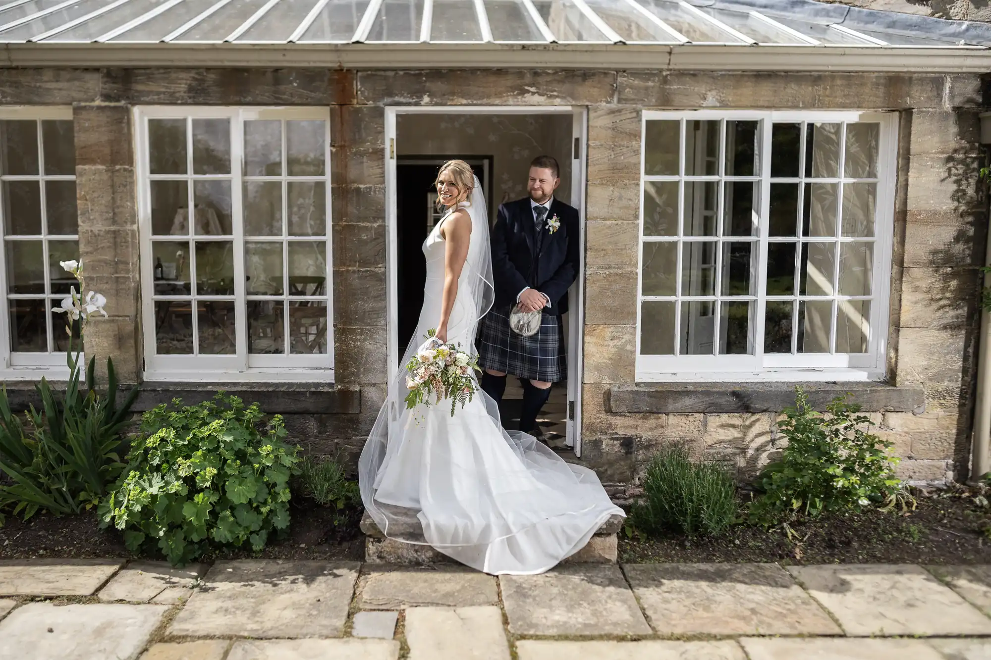 Bride in white dress holding a bouquet and groom in kilt standing by an open door of a stone building with large windows.