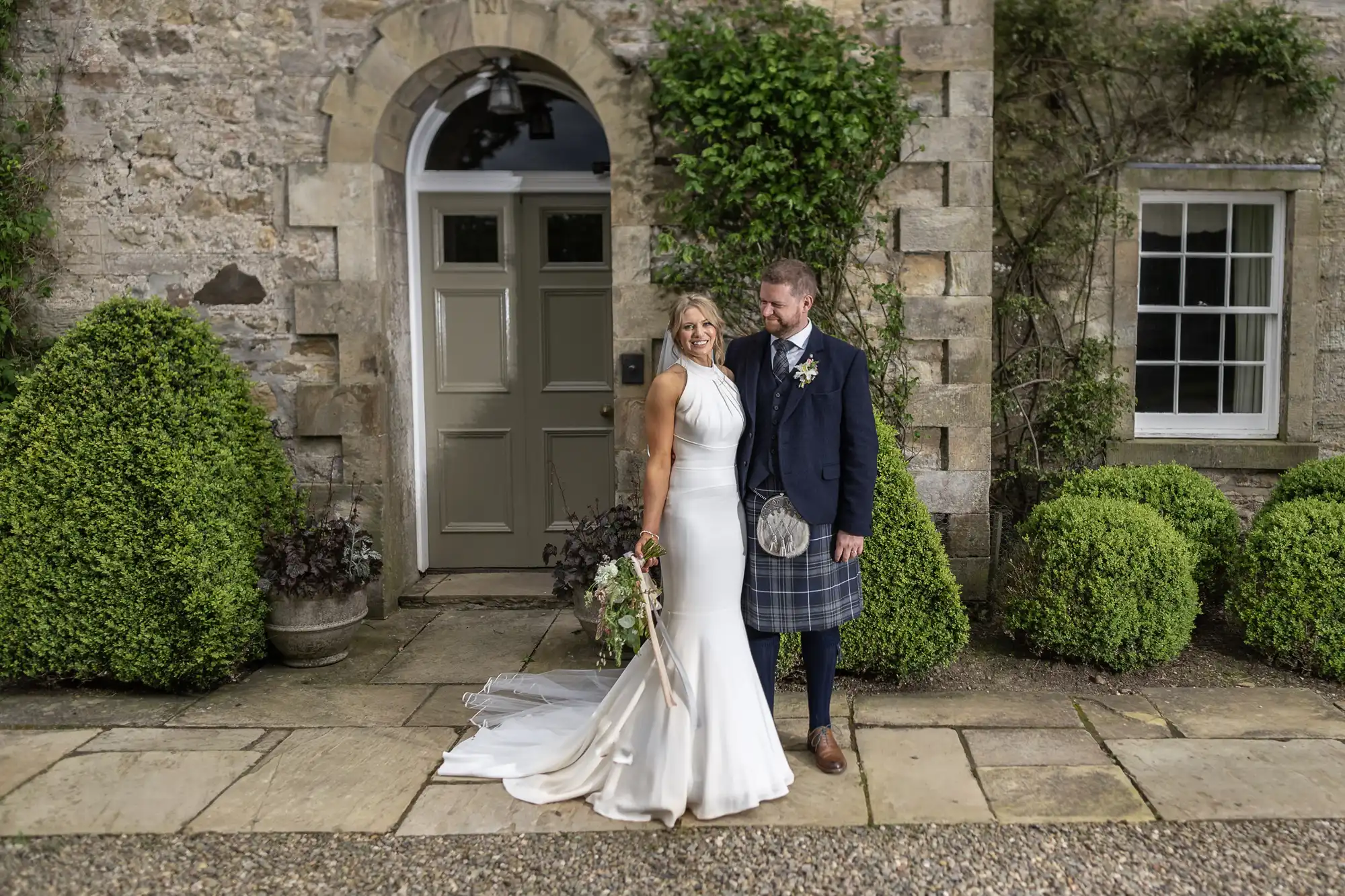 A bride in a white dress and a groom in a kilt stand smiling together outside a stone building flanked by shrubs.