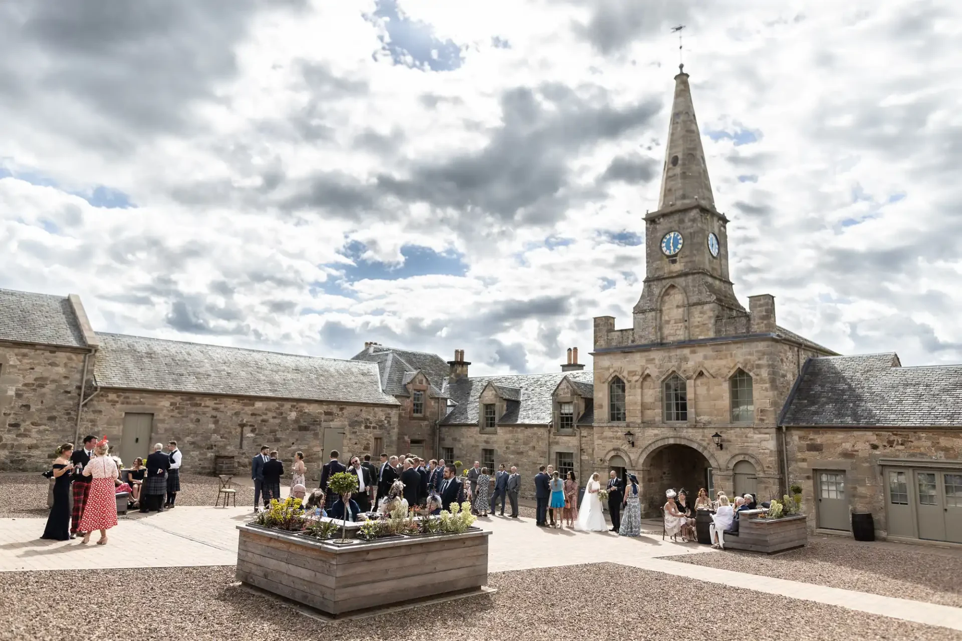 Guests gather at a spacious courtyard with historical stone buildings and a central clock tower under a partly cloudy sky at Rosebery Steading.