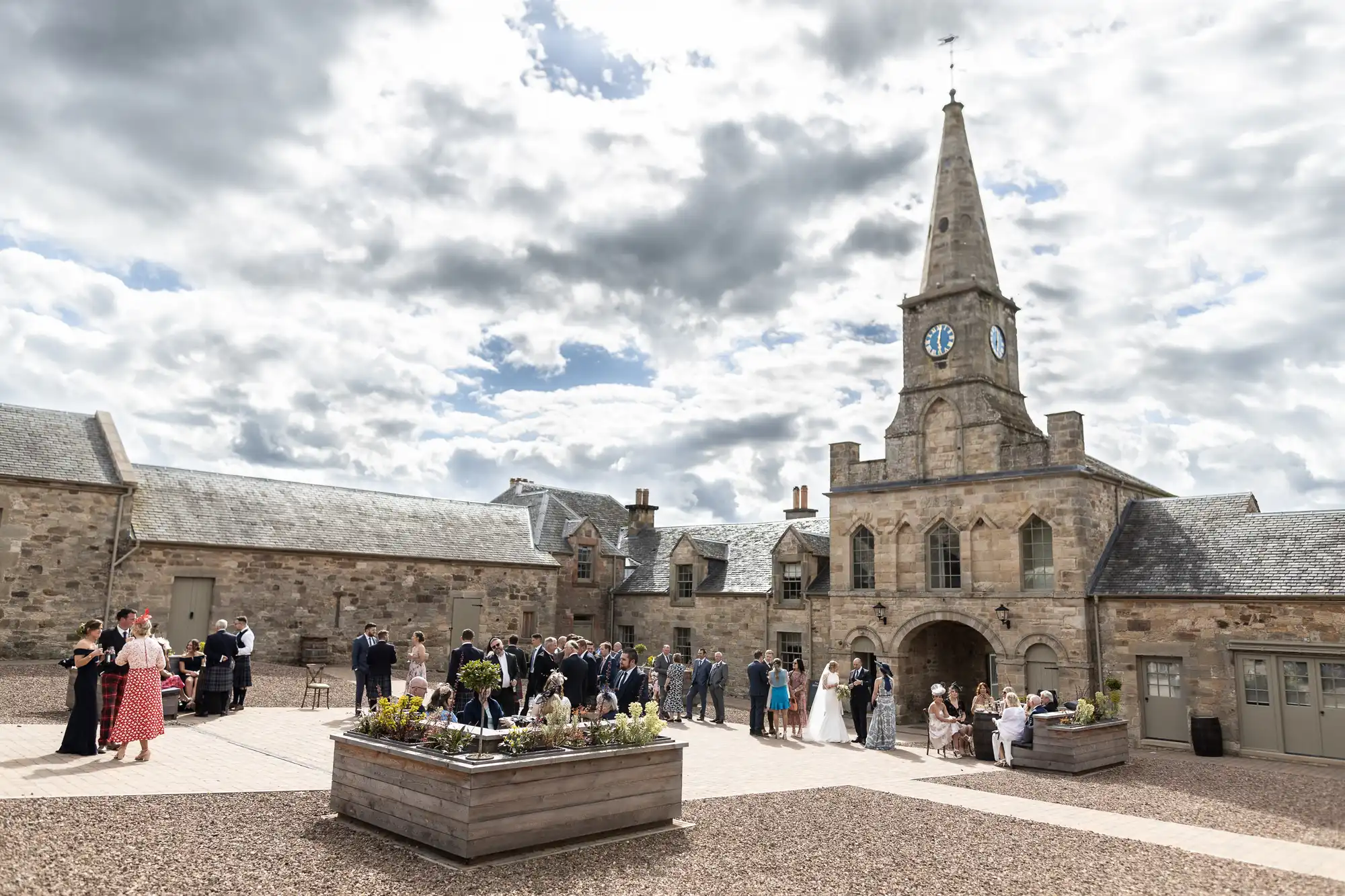 Guests gather at a spacious courtyard with historical stone buildings and a central clock tower under a partly cloudy sky.