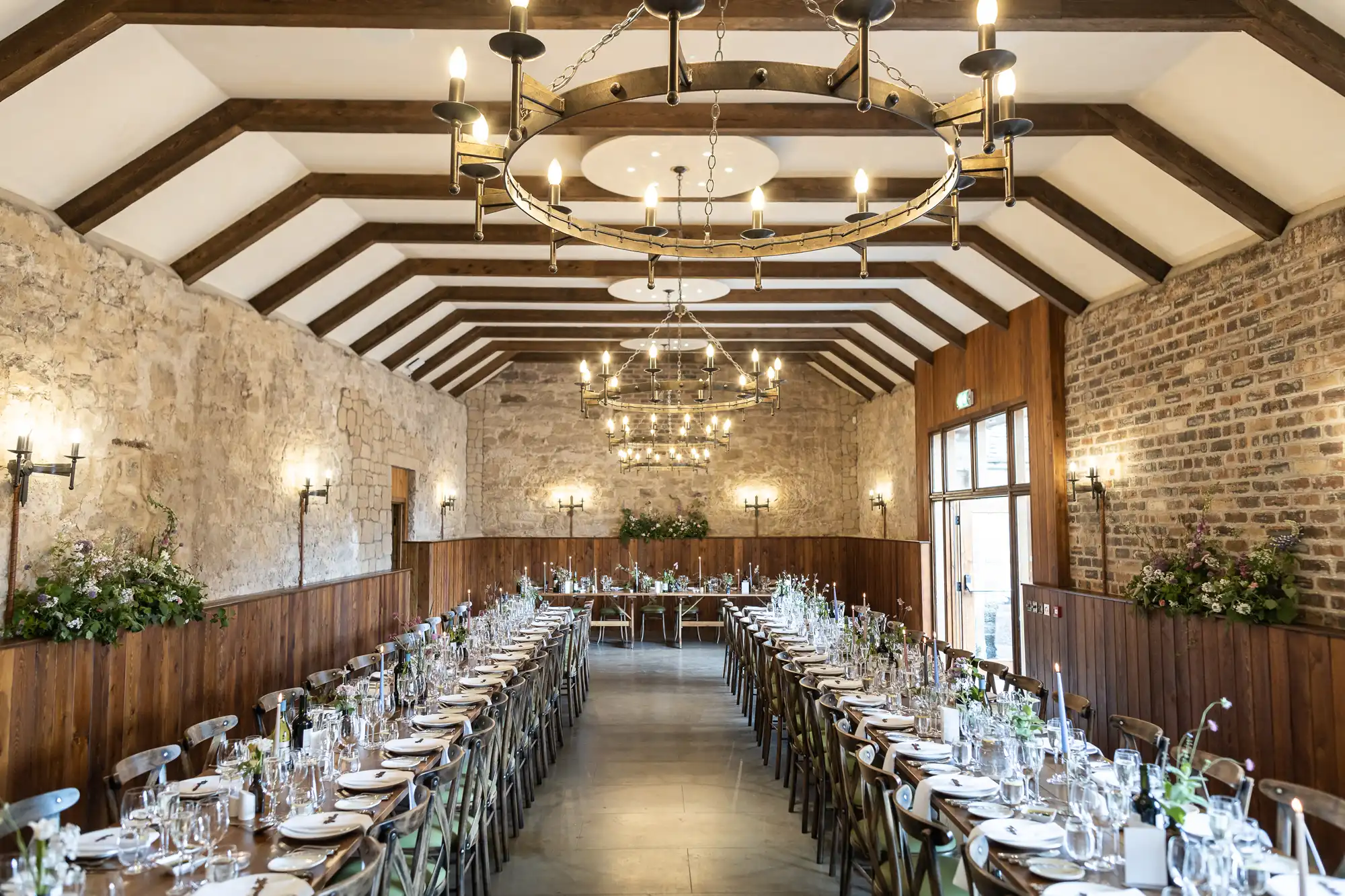 Elegant dining hall with exposed brick walls and wooden beams, featuring long tables set for a meal, adorned with candles and floral arrangements.