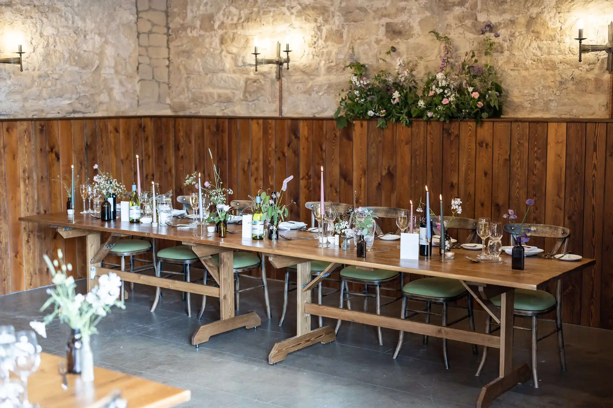 Rustic dining setup inside a stone-walled room with a long wooden table adorned with flowers, glasses, and bottles, flanked by green chairs.