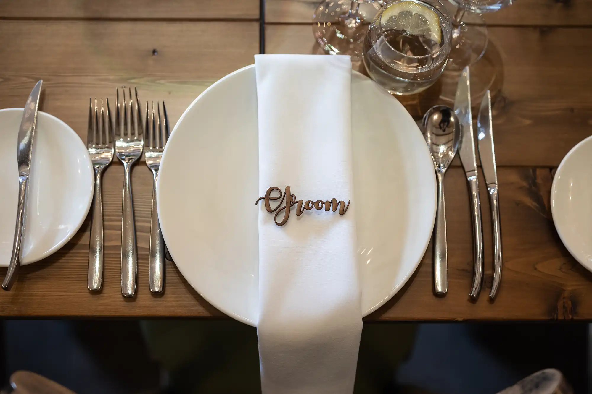 Elegant wedding table setting with a "Groom" place setting marker on a folded white napkin, surrounded by silverware and a glass on a wooden table.