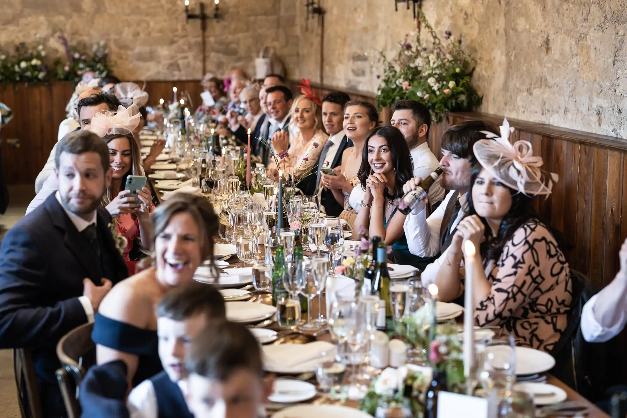 Guests at a long banquet table in a rustic setting, dressed in formal attire and interacting joyfully.