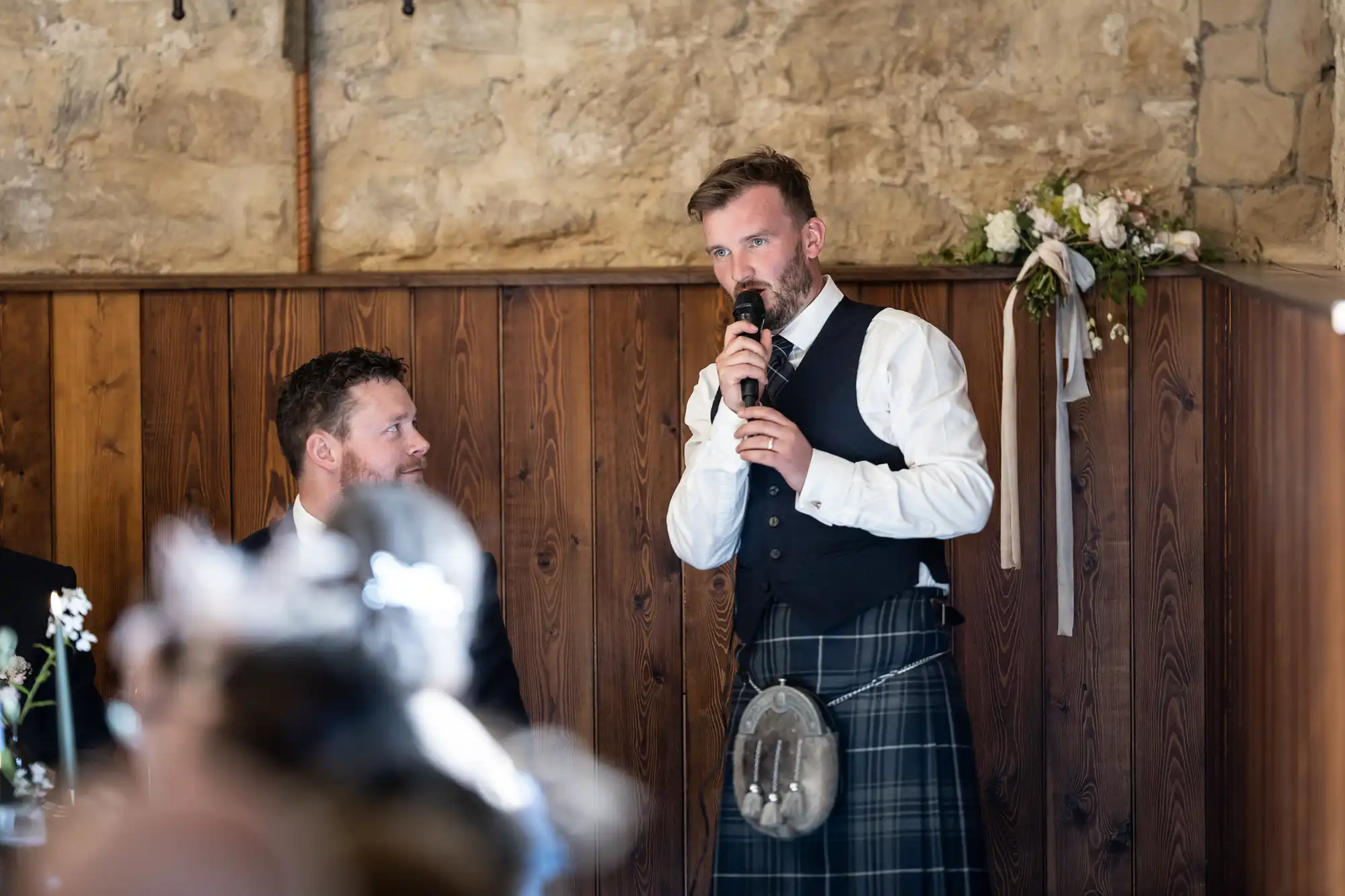 Man in a kilt giving a speech at a wedding, holding a microphone, with another man seated nearby, in a rustic stone and wood-paneled room.