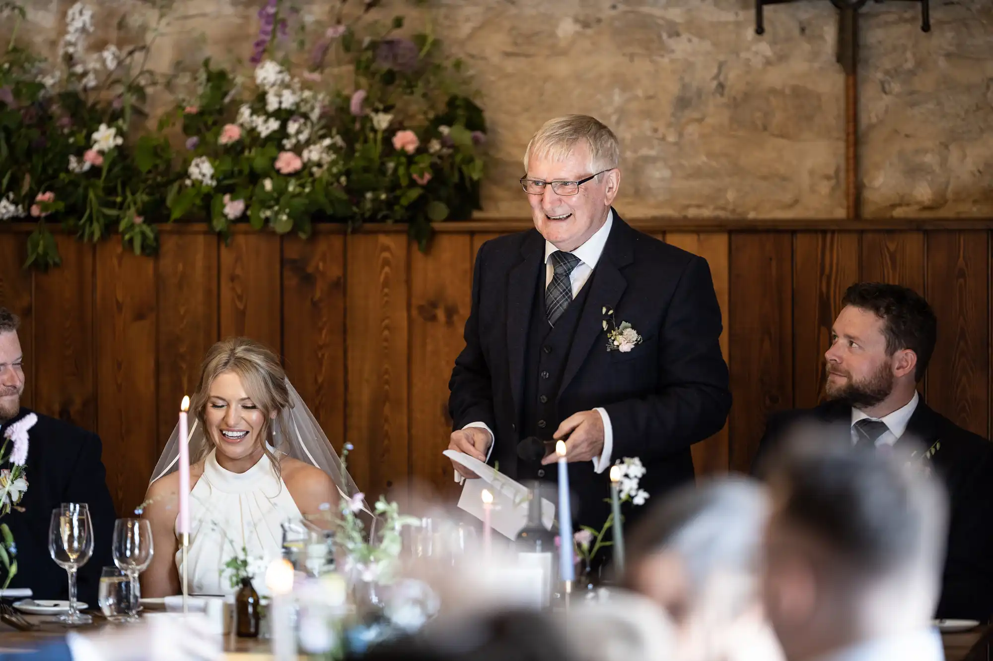 An elderly man gives a speech at a wedding reception, smiling guests including a bride and a groom seated at the table.