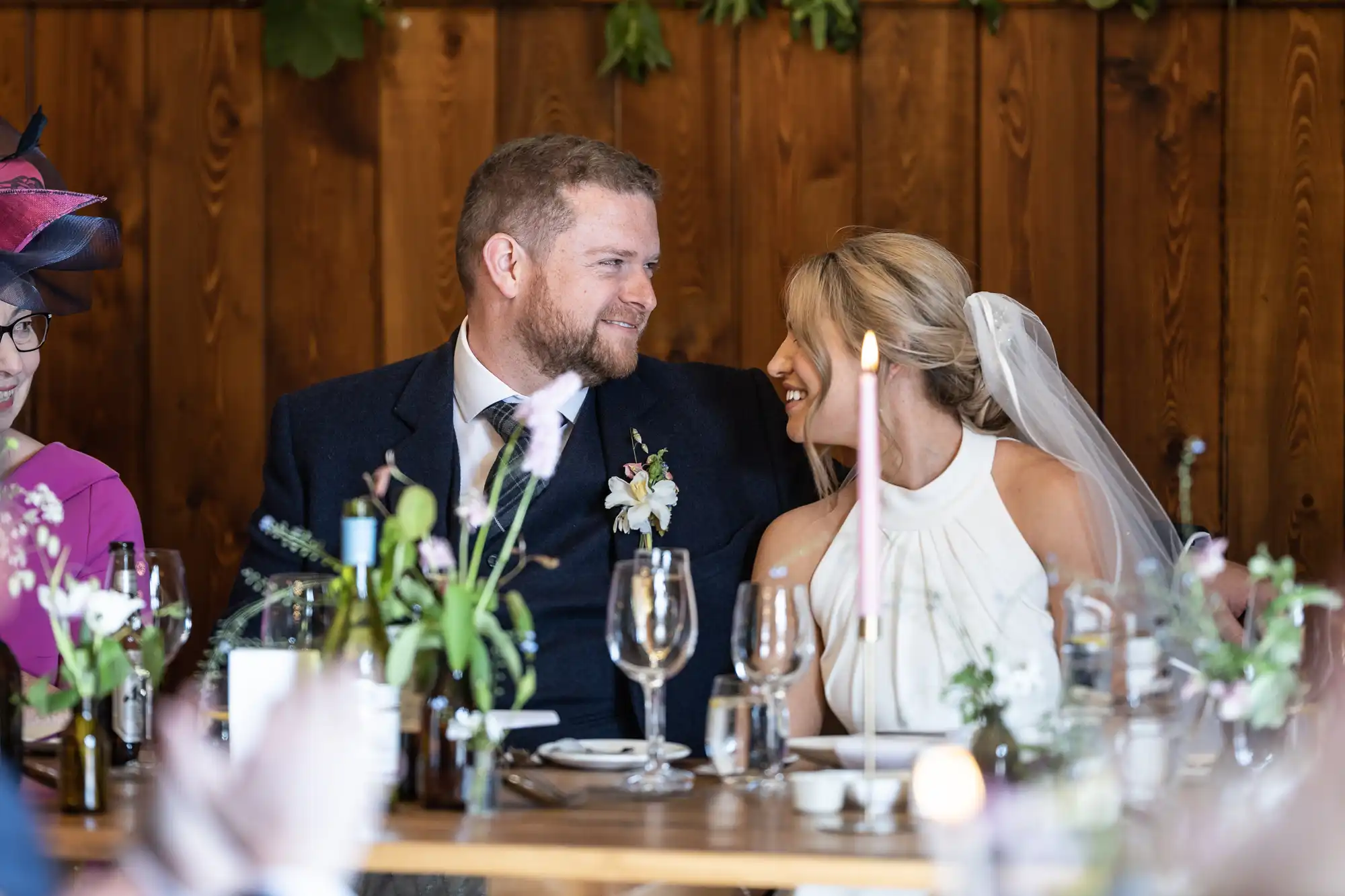 A bride and groom smiling at each other at their wedding reception table, surrounded by candles and floral decorations.