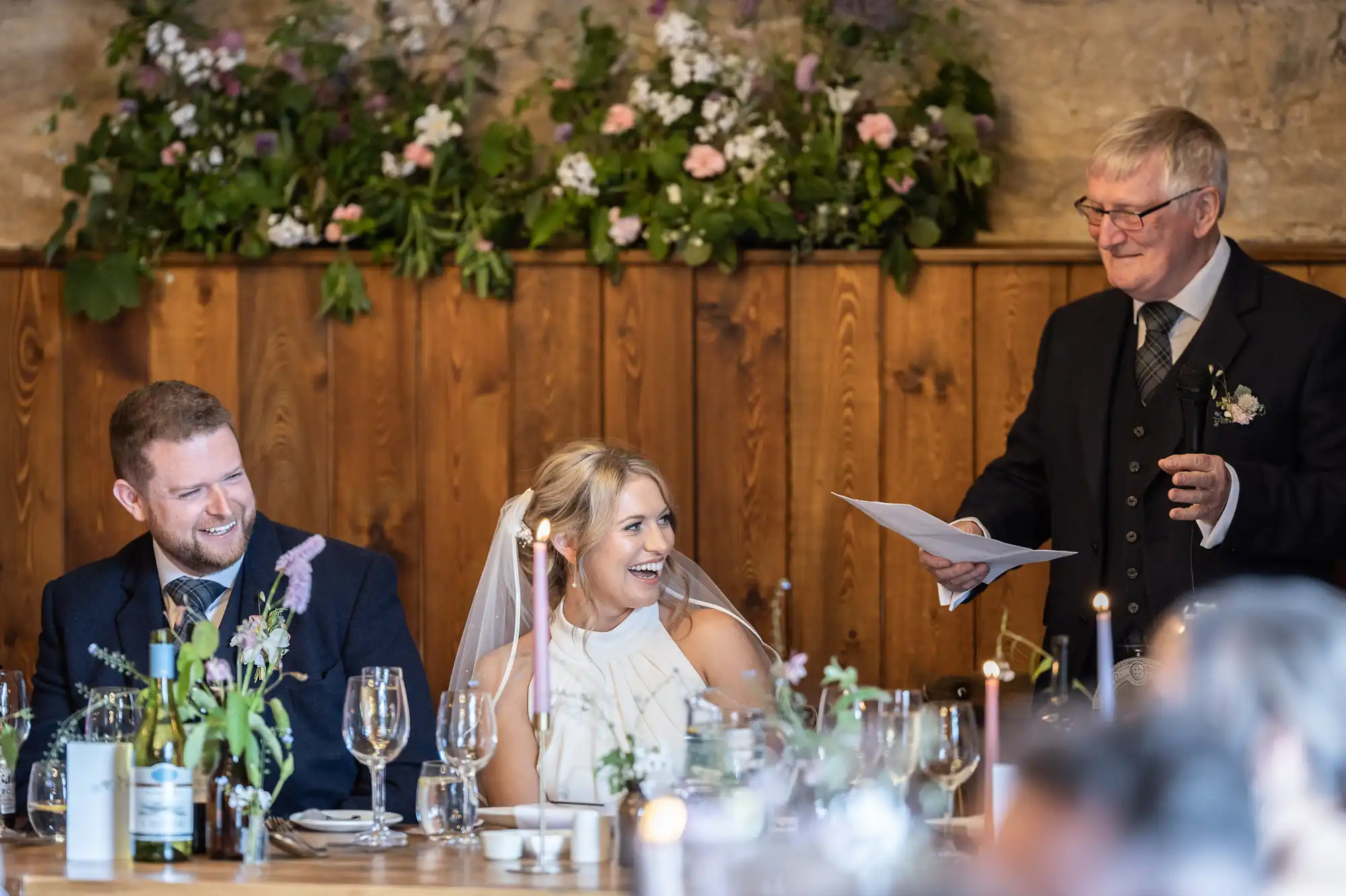 A bride and groom laugh at a speech given by an older man at their wedding reception, surrounded by floral decorations and guests.