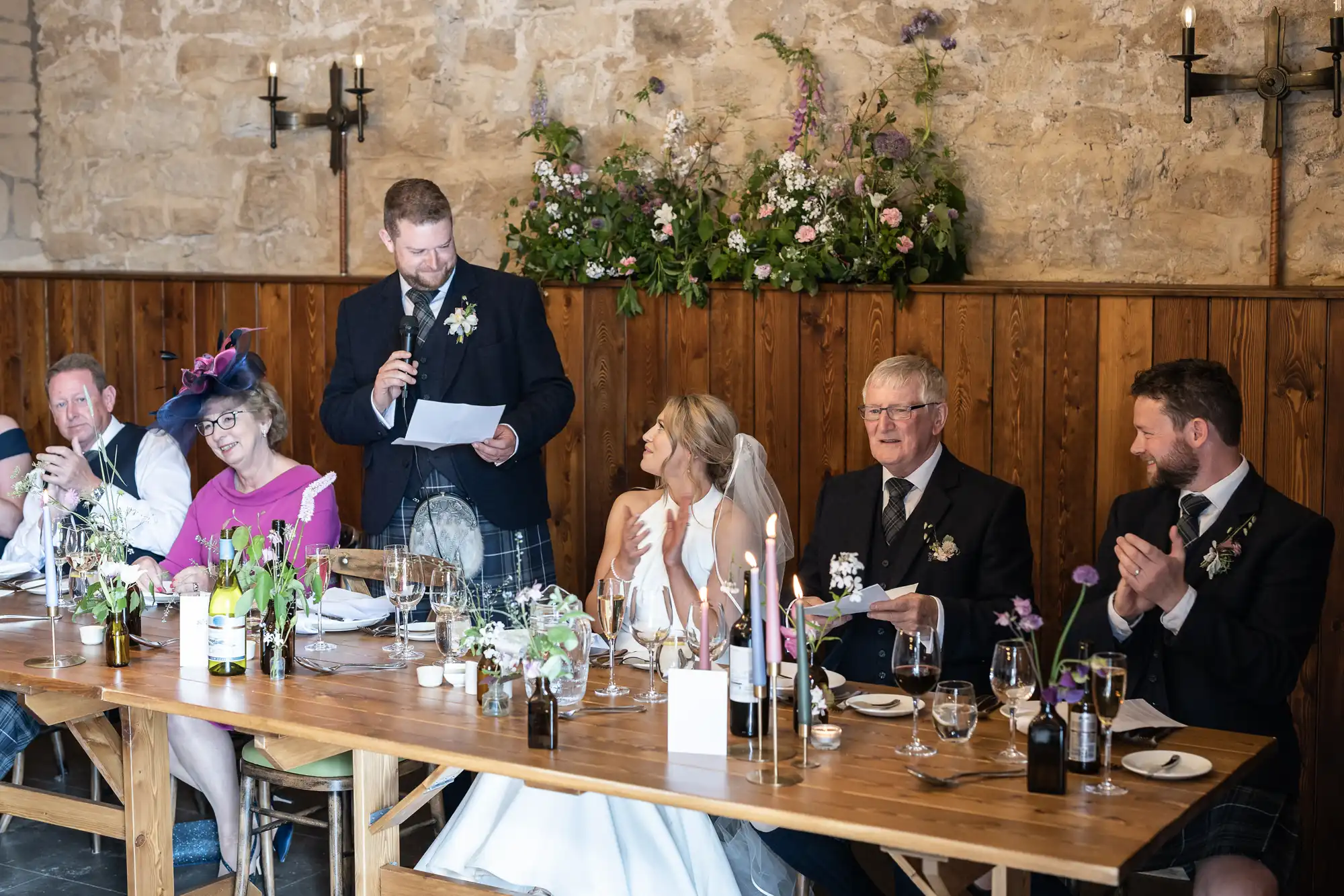 A man delivers a speech at a wedding reception table while other guests listen attentively in a rustic venue decorated with flowers.