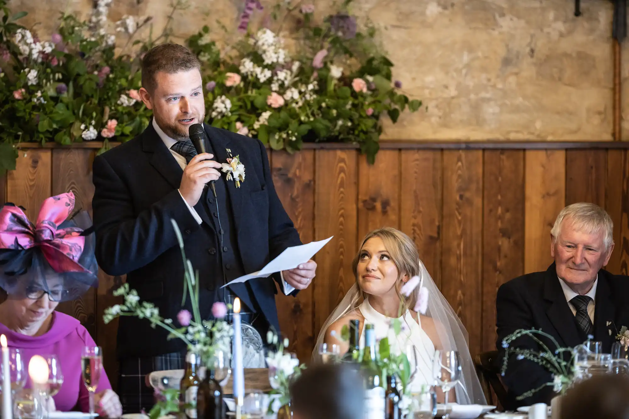 Man giving a speech at a wedding reception with a bride listening intently and other guests around a decorated table.