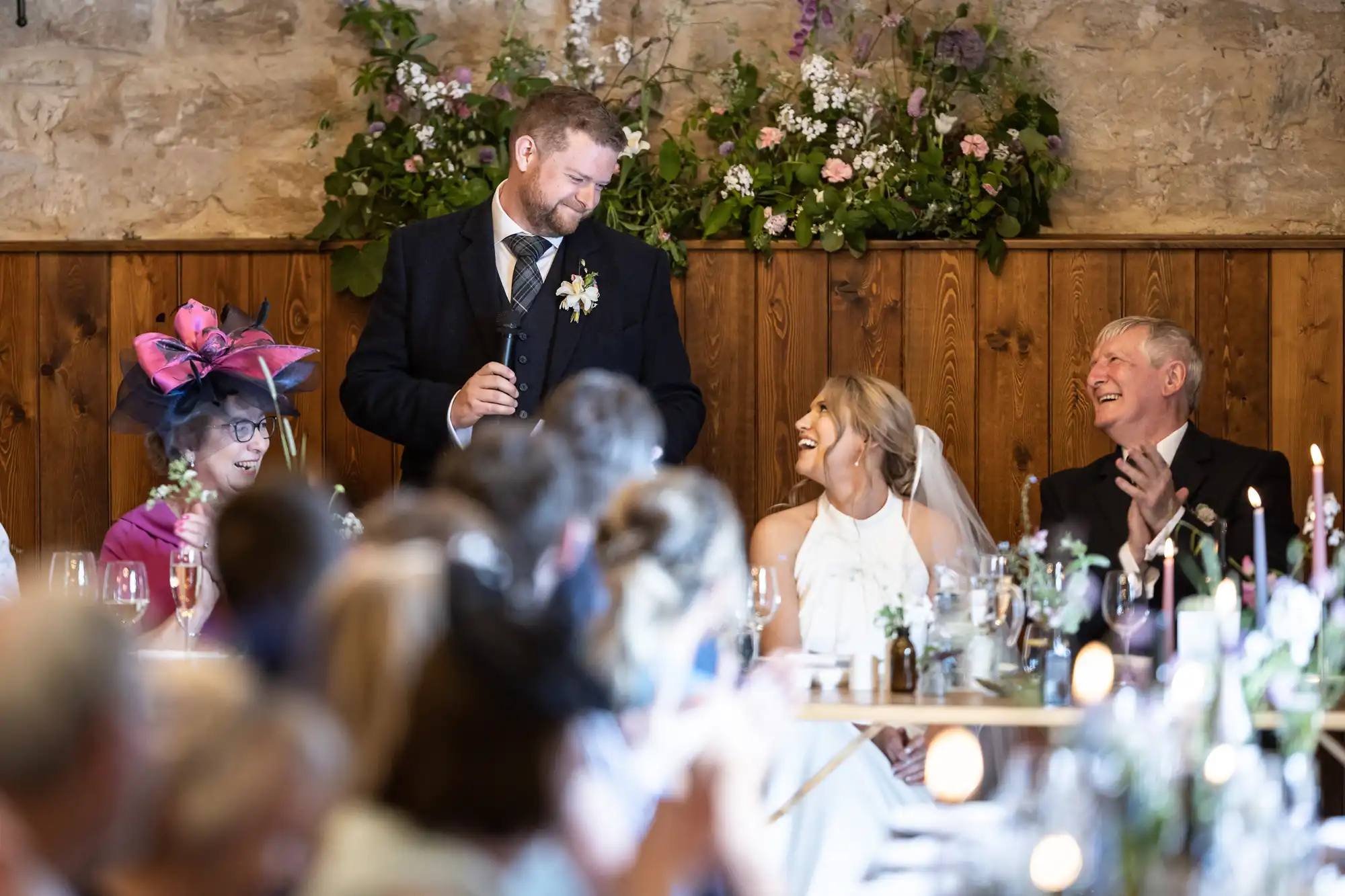 A groom giving a speech at a wedding reception, smiling at the bride, with guests laughing and enjoying the moment.