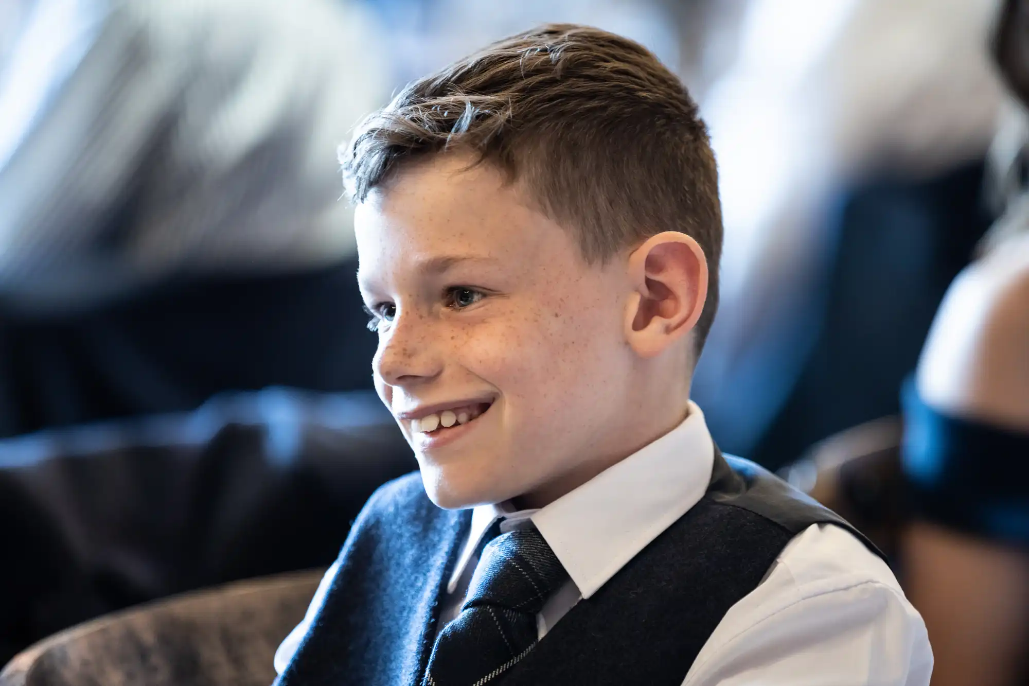 A young boy with freckles, smiling, dressed in a formal vest and tie at an indoor event.