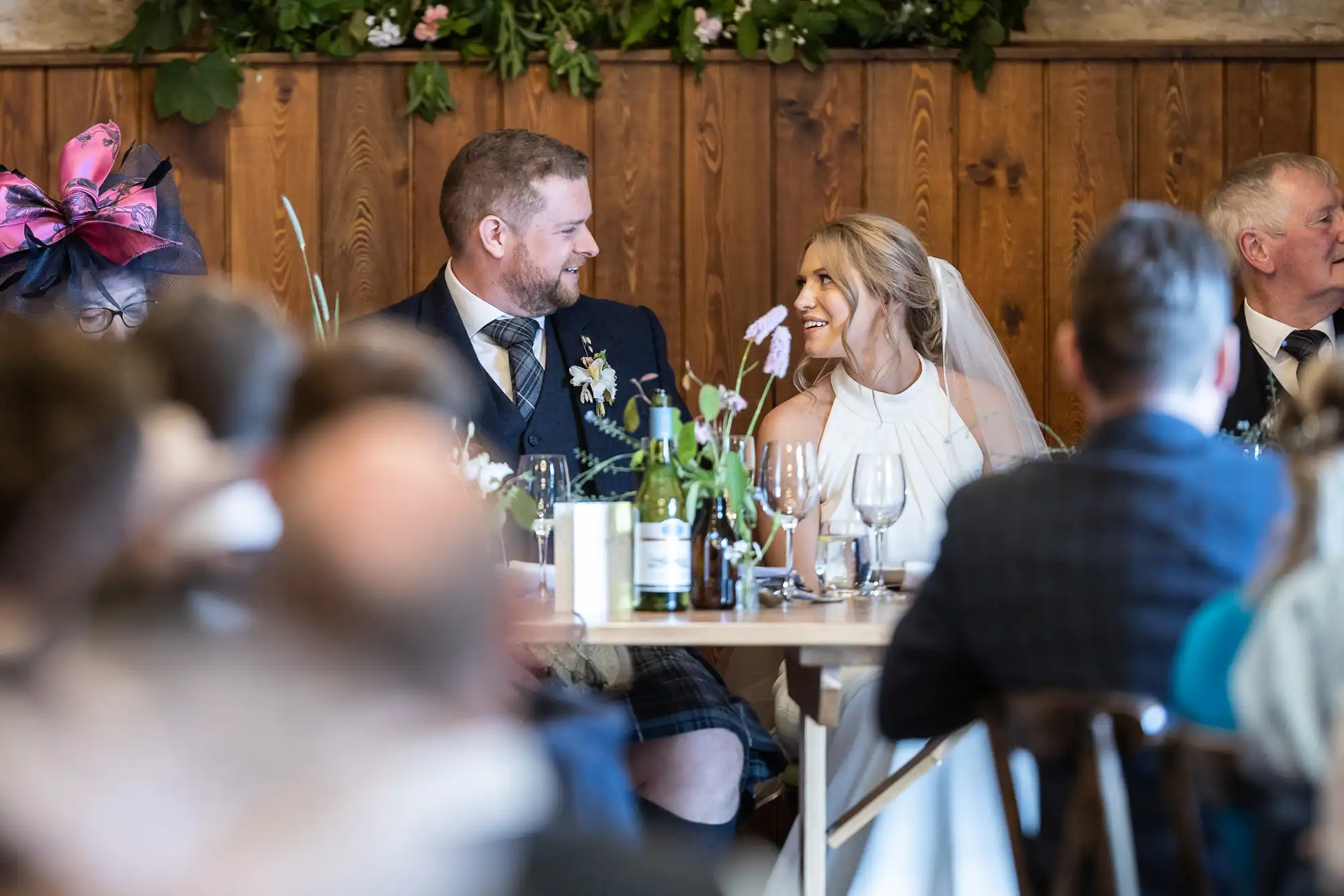 A bride and groom smiling at each other at a wedding reception table, surrounded by guests and rustic decor.