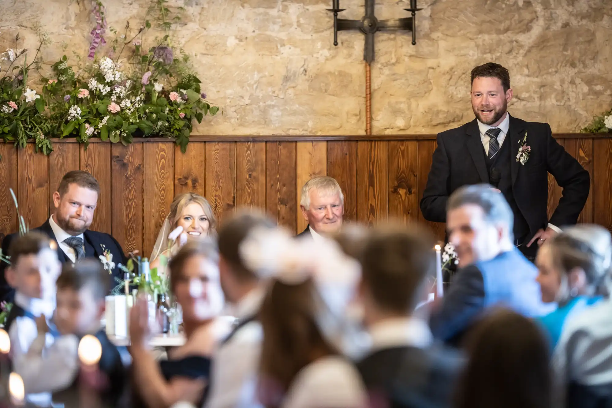 A man stands smiling at a wedding reception, with seated guests focused on him, in a rustic hall with flower arrangements.