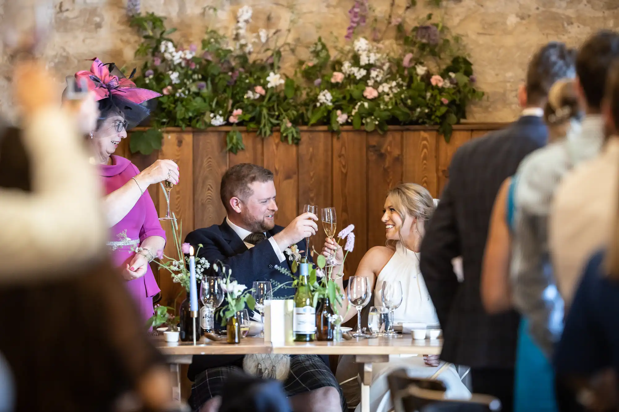 A bride and groom seated at a wedding reception table, smiling and toasting, with guests and floral decorations around them.