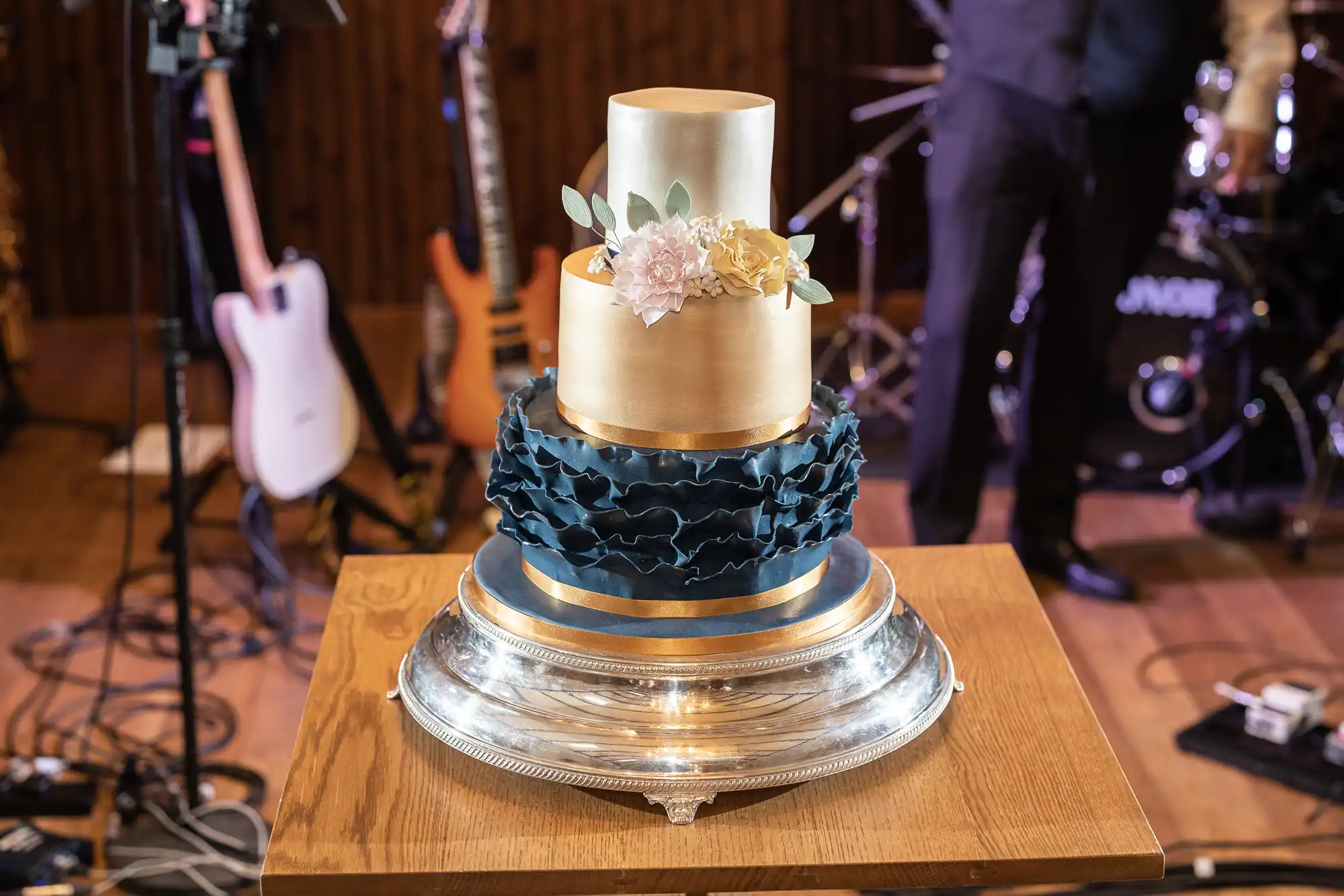 A three-tiered wedding cake adorned with gold and navy layers, accented with pink flowers, displayed on a silver stand with musical instruments in the background.