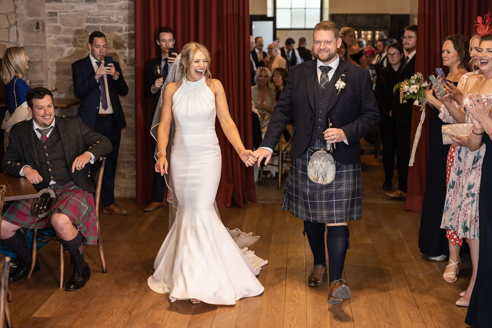 A bride in a white dress and a groom in a kilt smiling as they walk hand-in-hand through a crowd of cheering guests at a wedding venue.