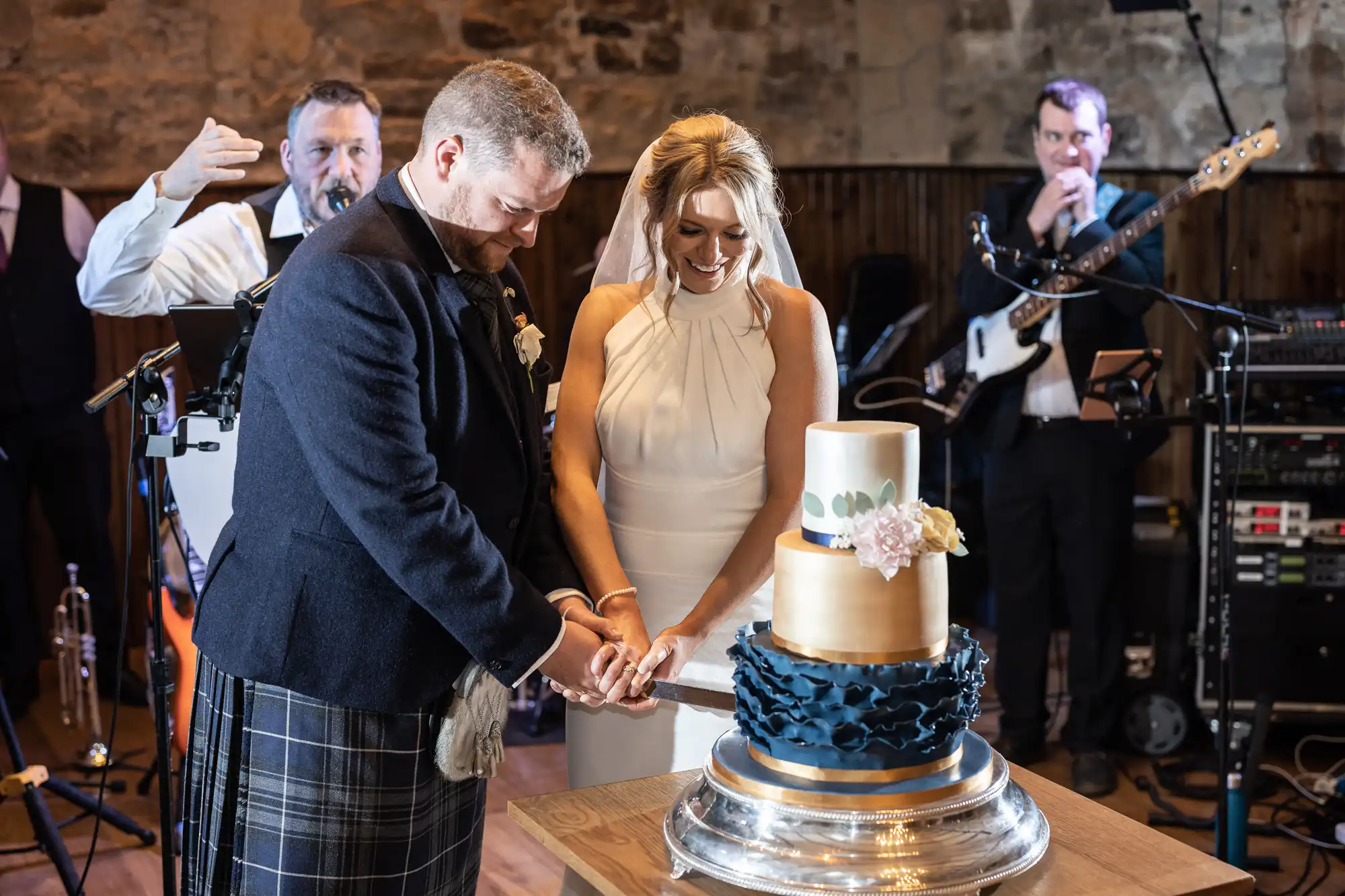 A bride and groom in a kilts cutting a tiered wedding cake, with a band playing in the background at a festive venue.