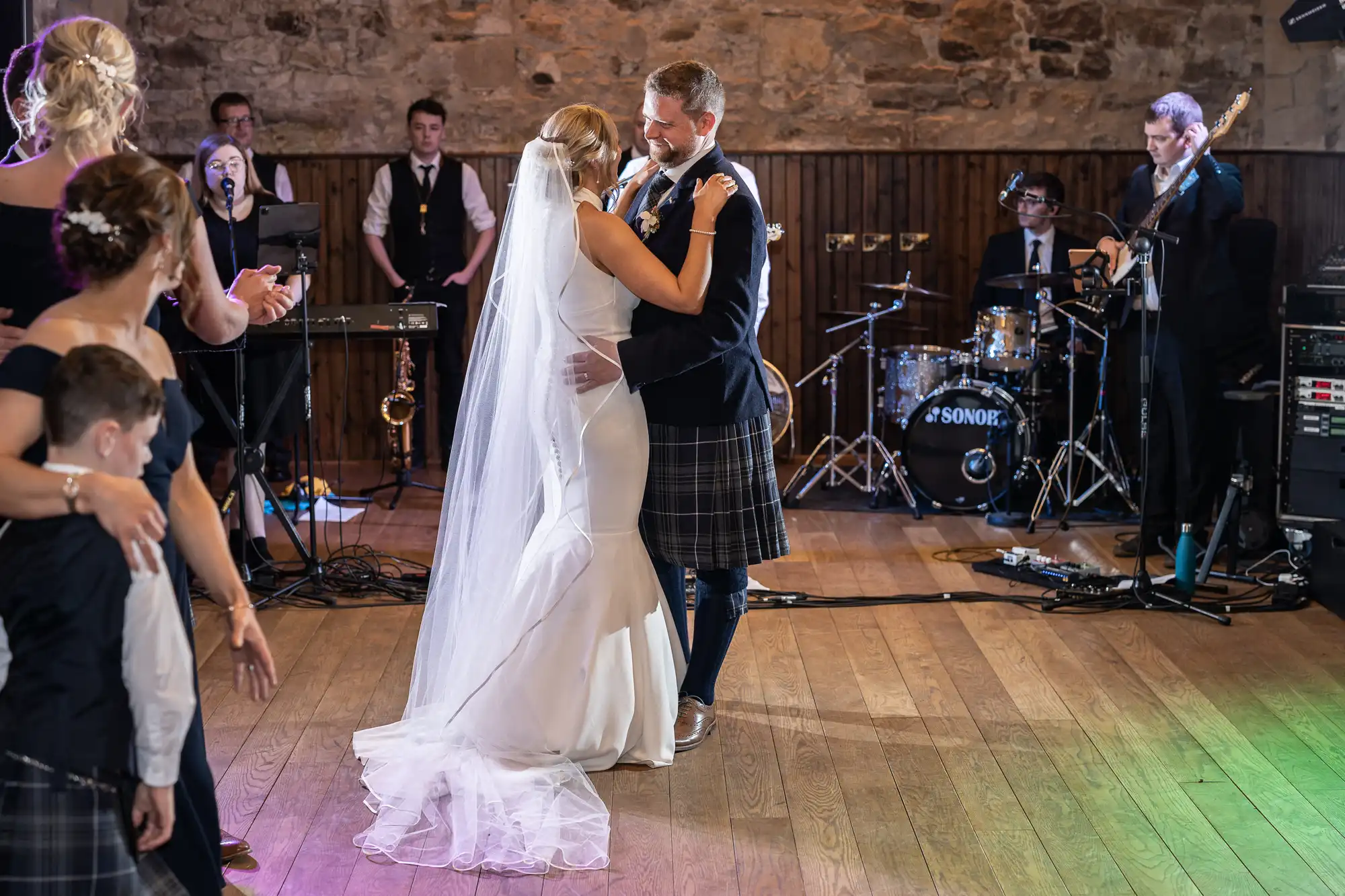 A bride and groom share their first dance in a hall with a live band and guests surrounding them. The groom wears a kilt, and the bride is in a white dress.