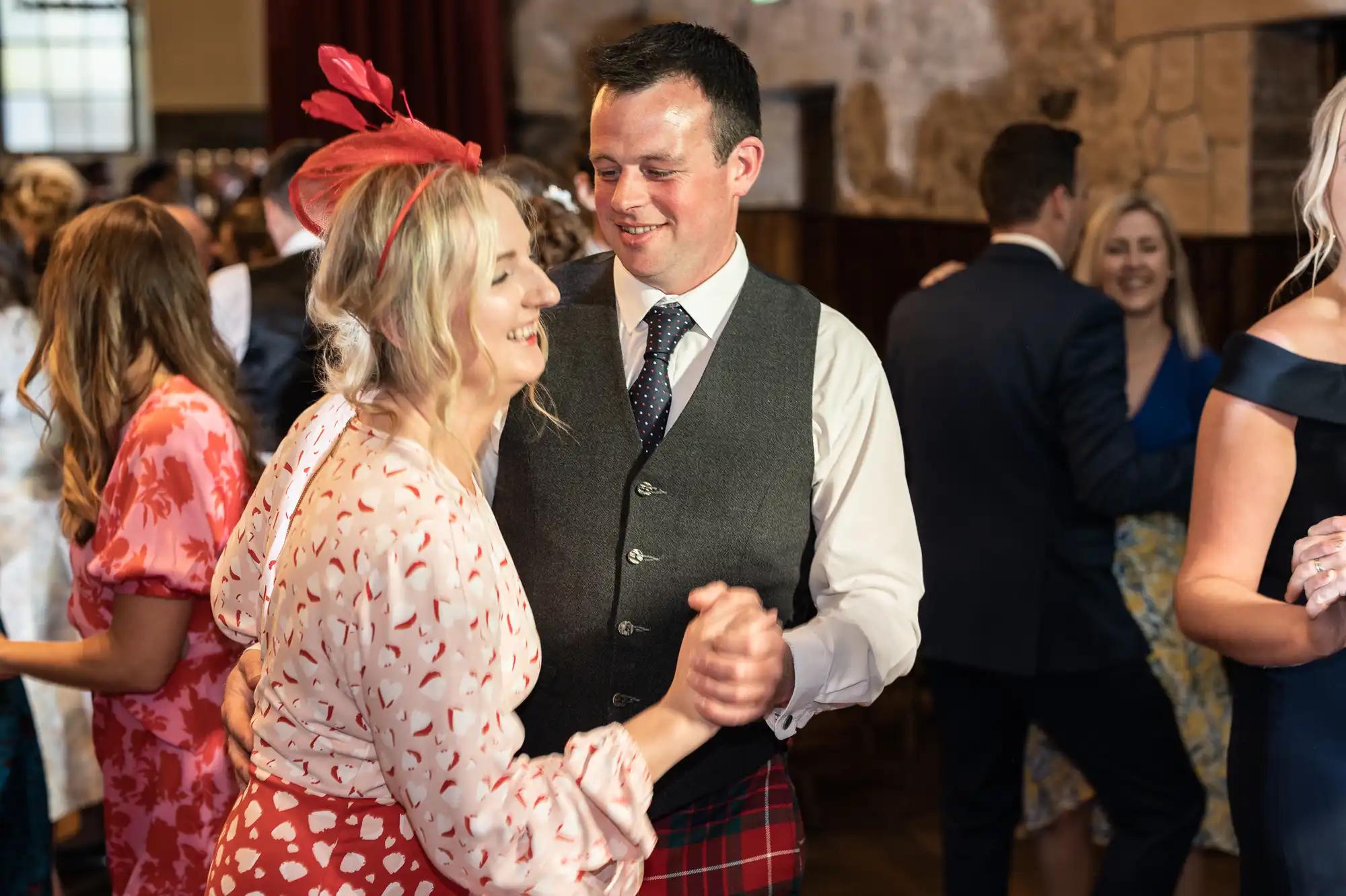 A man in a kilt and waistcoat dances with a smiling woman in a red and white dress at a lively indoor event.