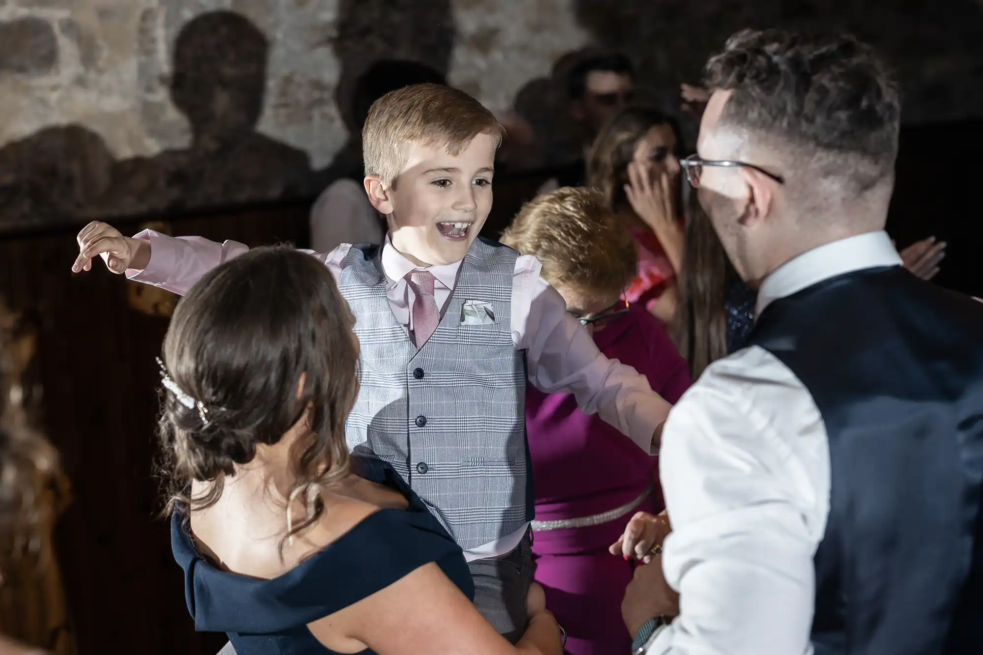 A young boy in a vest and tie joyfully dances at a party, pointing towards a man, with other guests around them enjoying the festivities.