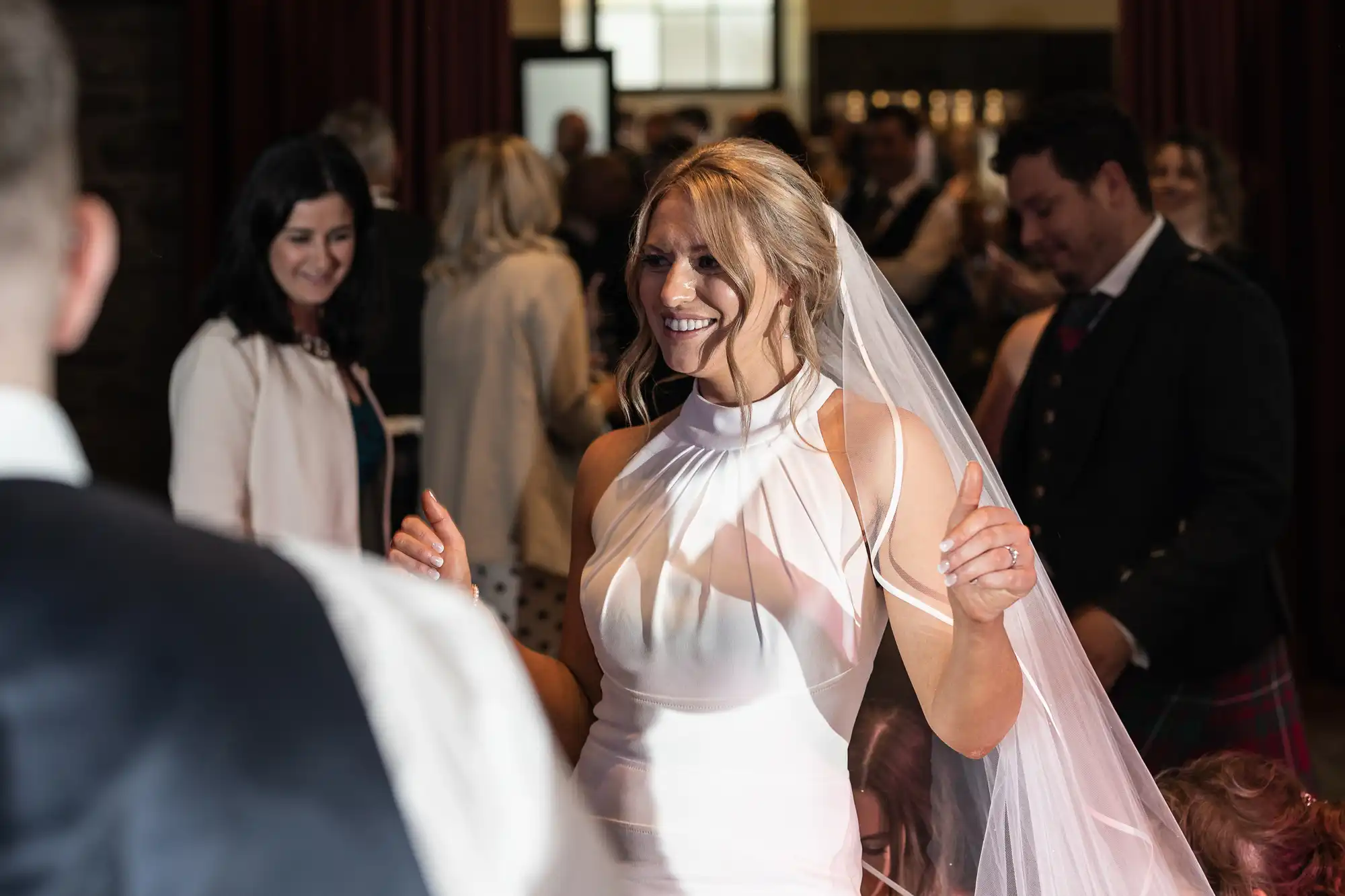 A bride in a white dress smiles joyfully at a guest during an indoor wedding reception, with blurred attendees in the background.