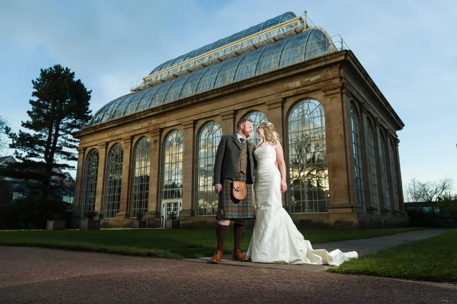 Royal Botanic Garden Edinburgh wedding photography - natural floral backdrop photos. A couple in wedding attire stands in front of a large, glass-domed building. The groom is in a kilt and jacket, while the bride wears a strapless white gown with a long train.