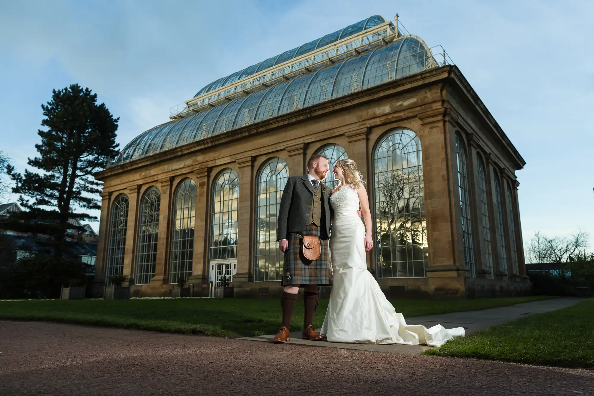 A couple in wedding attire stands in front of a large, glass-domed building. The groom is in a kilt and jacket, while the bride wears a strapless white gown with a long train.