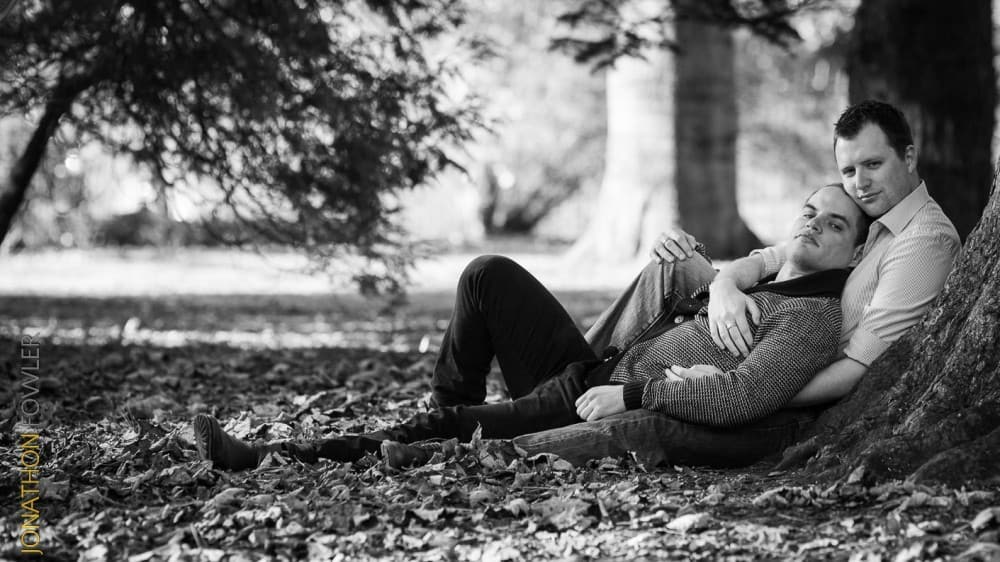 Royal Botanical Garden Edinburgh Pre-wedding photoshoot: Two men, one sitting and the other lying in his lap, pose under a large tree in a park. The scene is surrounded by fallen leaves with trees in the background. The photo is in black and white.