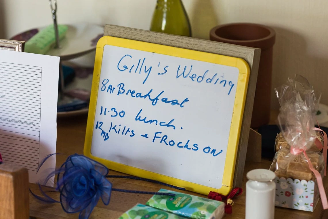 Handwritten schedule for "gilly's wedding" on a whiteboard among various items on a table, detailing breakfast and lunch times, and a note to put kilts and frocks on.