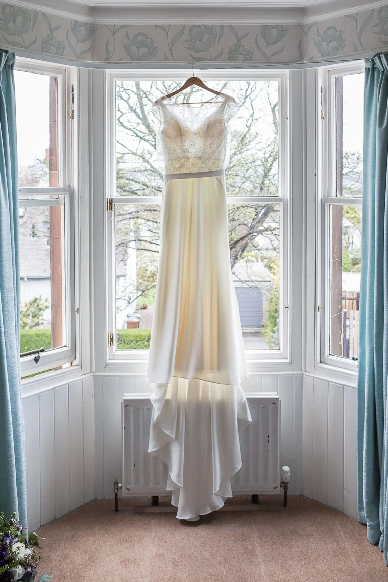 A wedding dress hanging in front of a bay window in a room with floral wallpaper and light blue curtains.