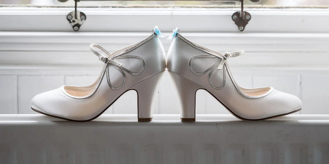 A pair of white bridal shoes with high heels and decorative straps placed on a white windowsill.