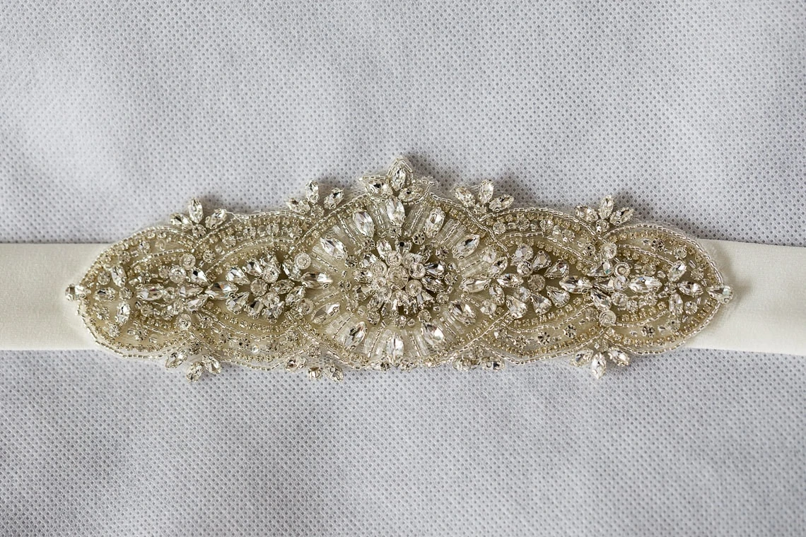 Embroidered bridal sash with intricate beadwork and crystals on a satin ribbon, displayed on a textured gray fabric.