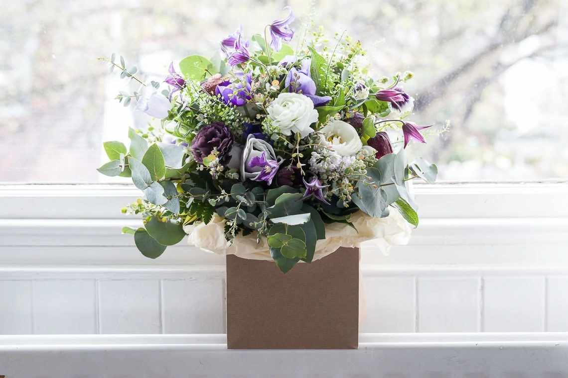 A vibrant bouquet of flowers including purple, white, and green blooms, positioned in a brown paper bag on a white windowsill.