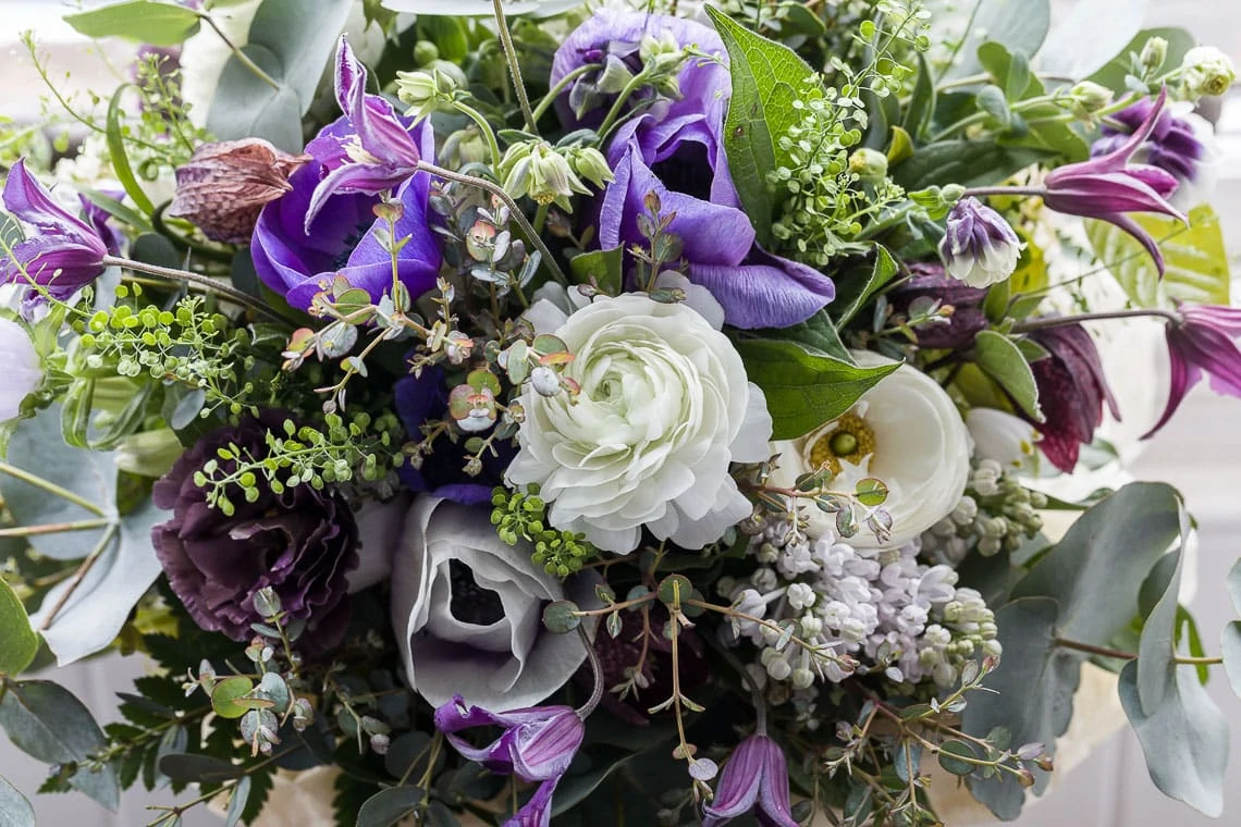 A close-up of a vibrant flower arrangement featuring purple, white, and green flowers, including lilacs and roses, interspersed with greenery.