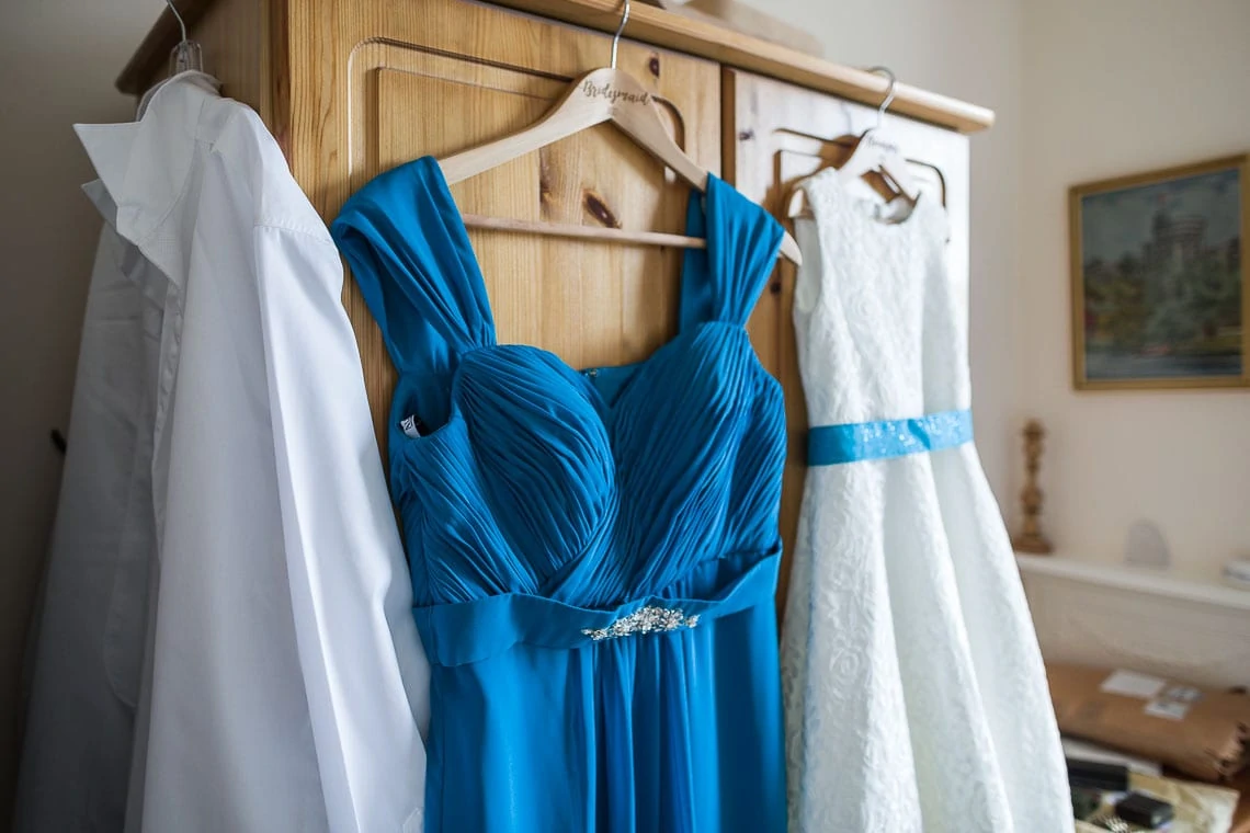A blue bridesmaid dress and a white wedding gown hanging on a wooden wardrobe in a room.