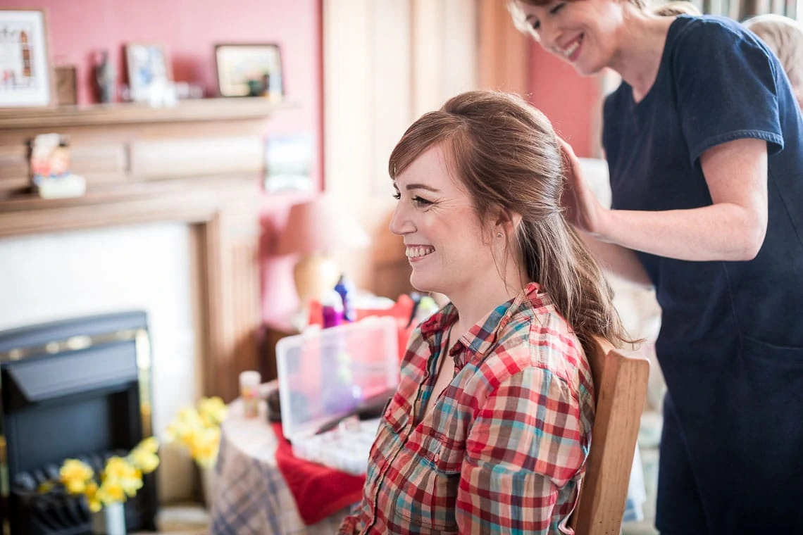A woman in a plaid shirt smiles while another woman styles her hair in a cozy living room setting.