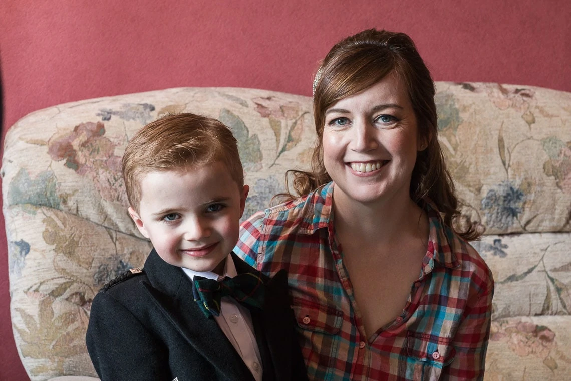 A smiling woman and a young boy in a tuxedo sitting together on a floral sofa, with a pink wall in the background.
