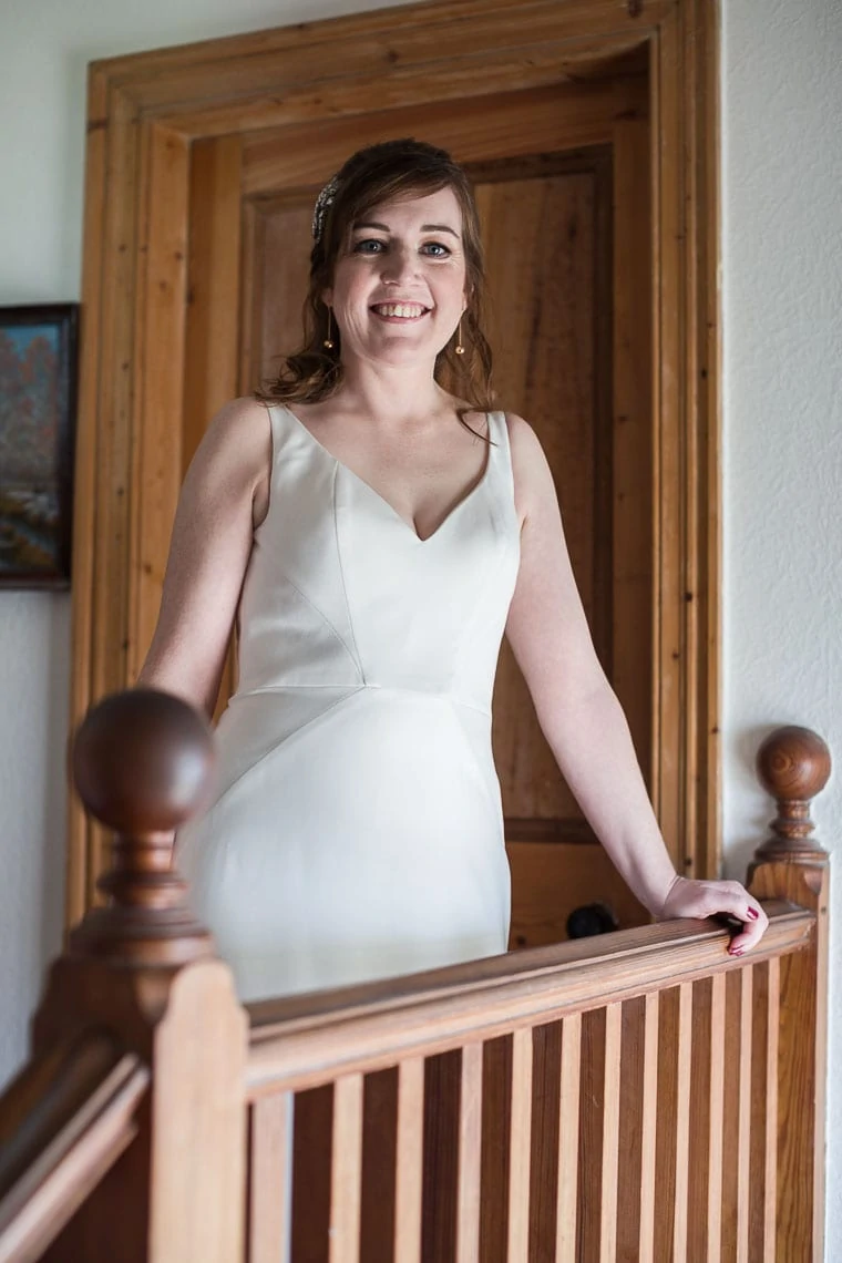 A smiling woman in a white dress standing at a wooden staircase in a home.