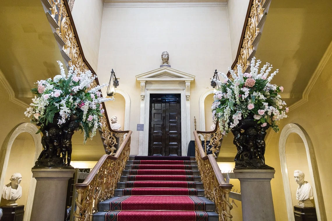 An elegant staircase with a red carpet flanked by two ornate floral arrangements, dark wood doors at the top, and busts on pedestals at the base.