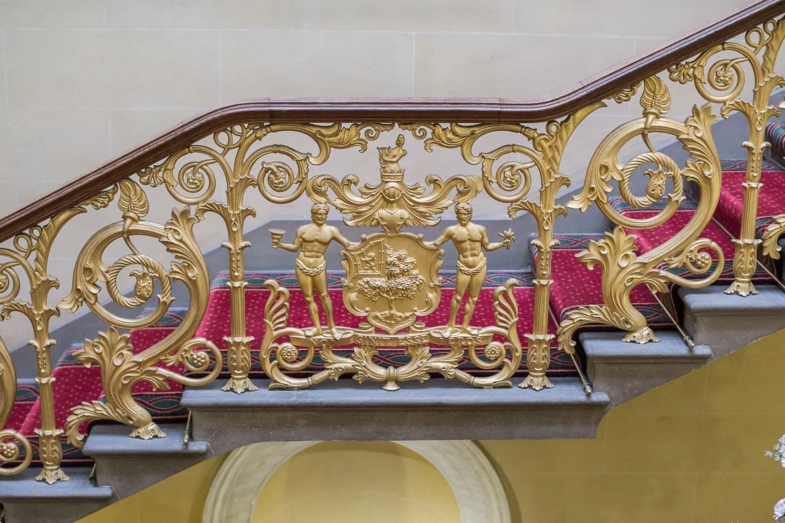 Ornate golden staircase railing with intricate designs and a central coat of arms flanked by sculpted figures, set against a marble stairway with red carpet.