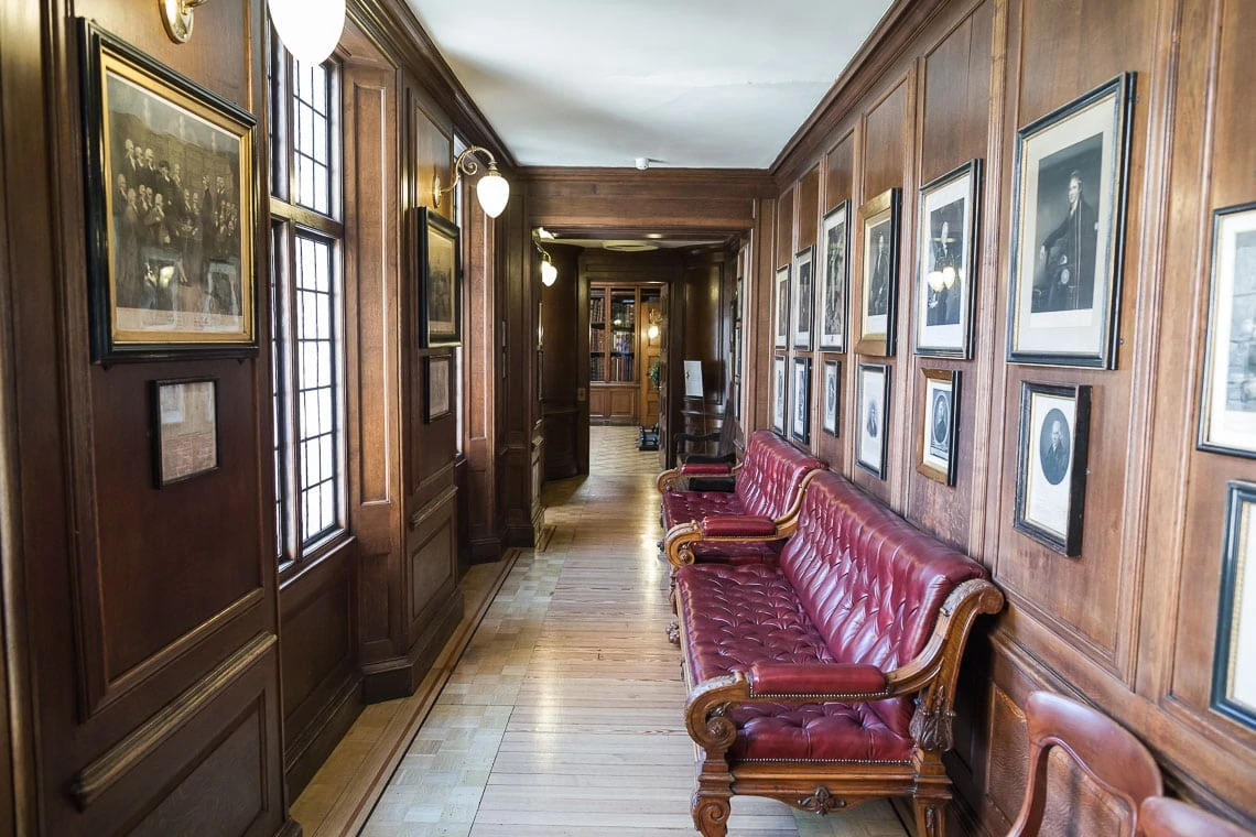 Narrow hallway lined with wood panels and historic portraits, featuring a red leather bench and hanging lamps.