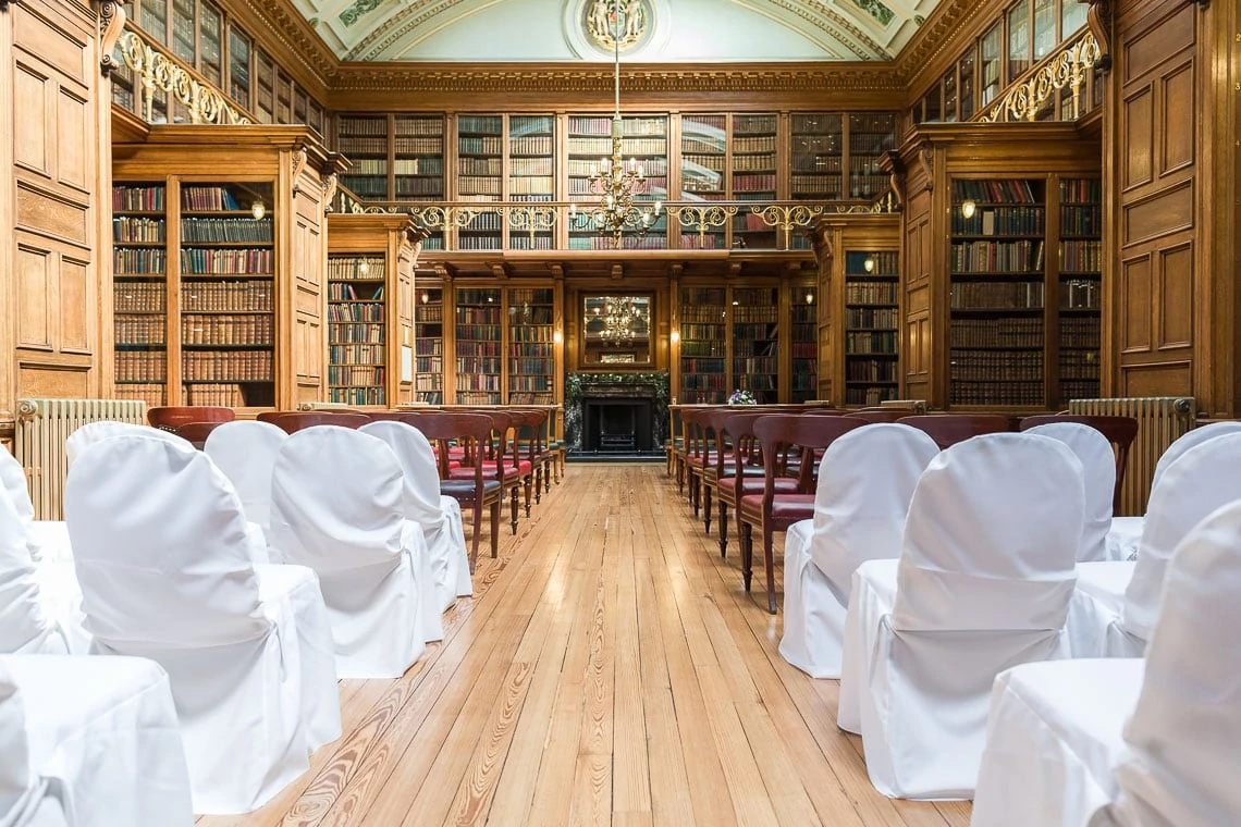 Interior of an ornate vintage library with rows of white chairs set up for an event, featuring wooden bookshelves filled with books and a central fireplace.