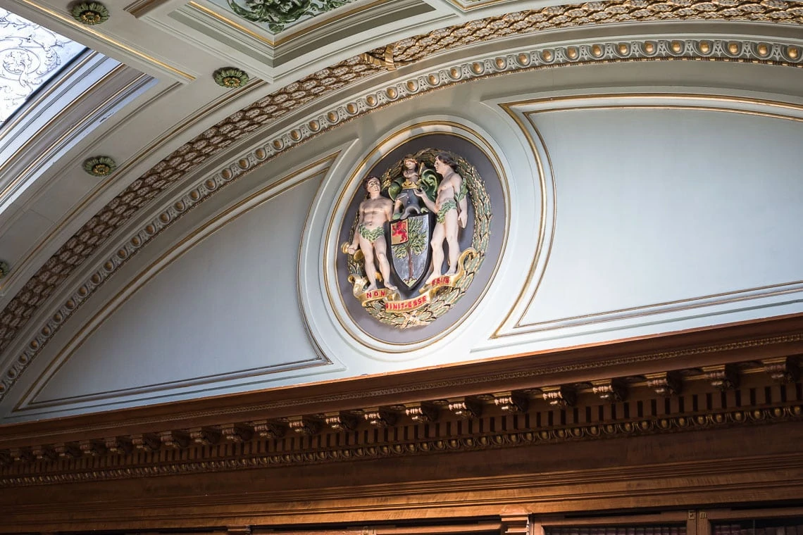 Ornate ceiling fresco featuring three classical figures within an intricate crest, surrounded by decorative gold and white mouldings in an elegant room.