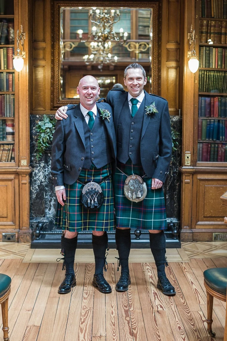 Two men in traditional scottish attire, including kilts and sporrans, standing and smiling in a library with wooden shelves and books.